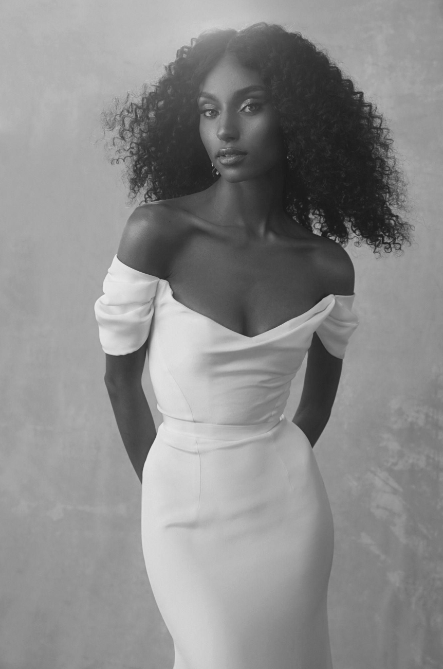 Black and white portrait of a woman with curly hair wearing an off-the-shoulder dress with puffed sleeves. She is looking at the camera with a serious expression, standing against a plain background.