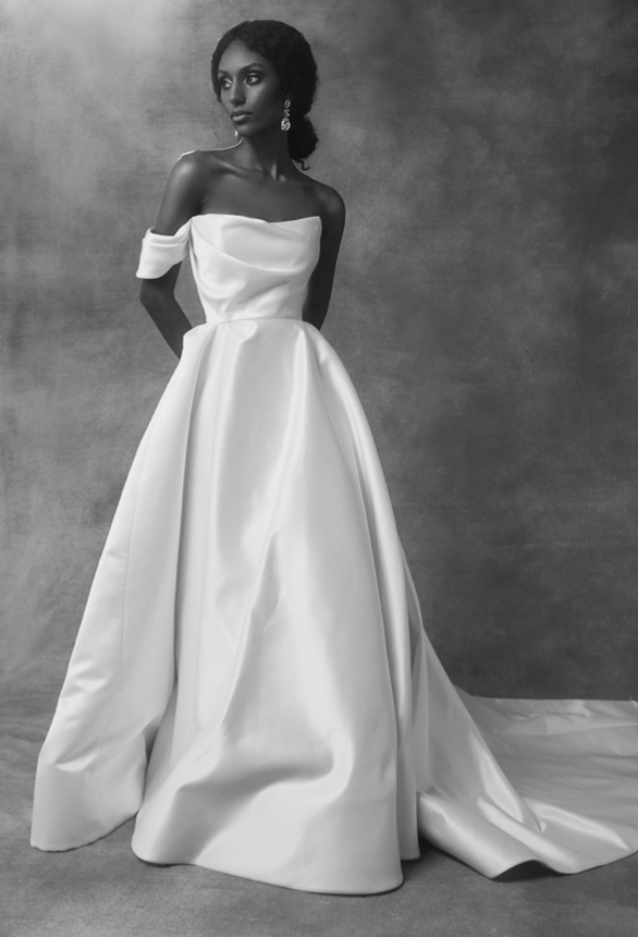 A woman in an off-the-shoulder white gown with a full skirt standing against a textured background.