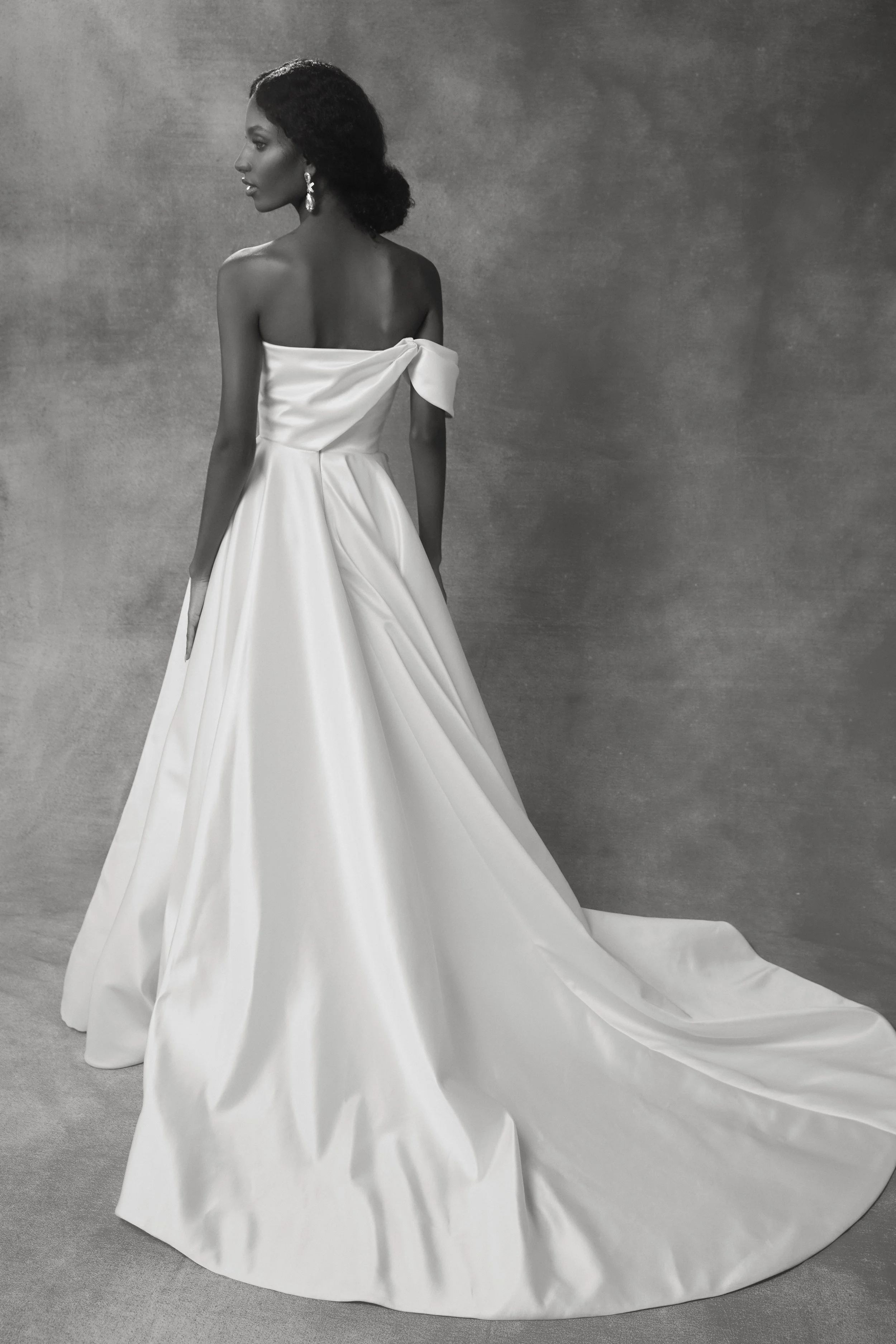A woman in a strapless white wedding gown with a long train, looking to her left, in front of a plain wall.
