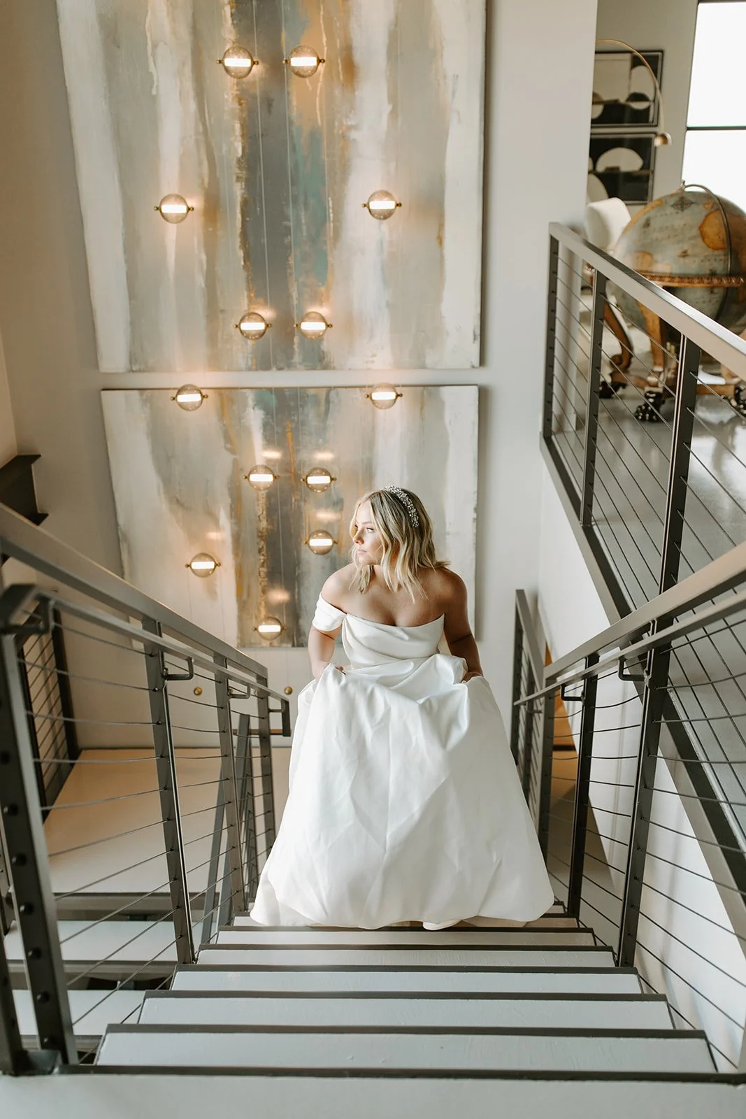 Brunette woman in an off-shoulder white wedding gown standing on a staircase, looking to the right, with a large abstract painting and modern light fixtures in the background.