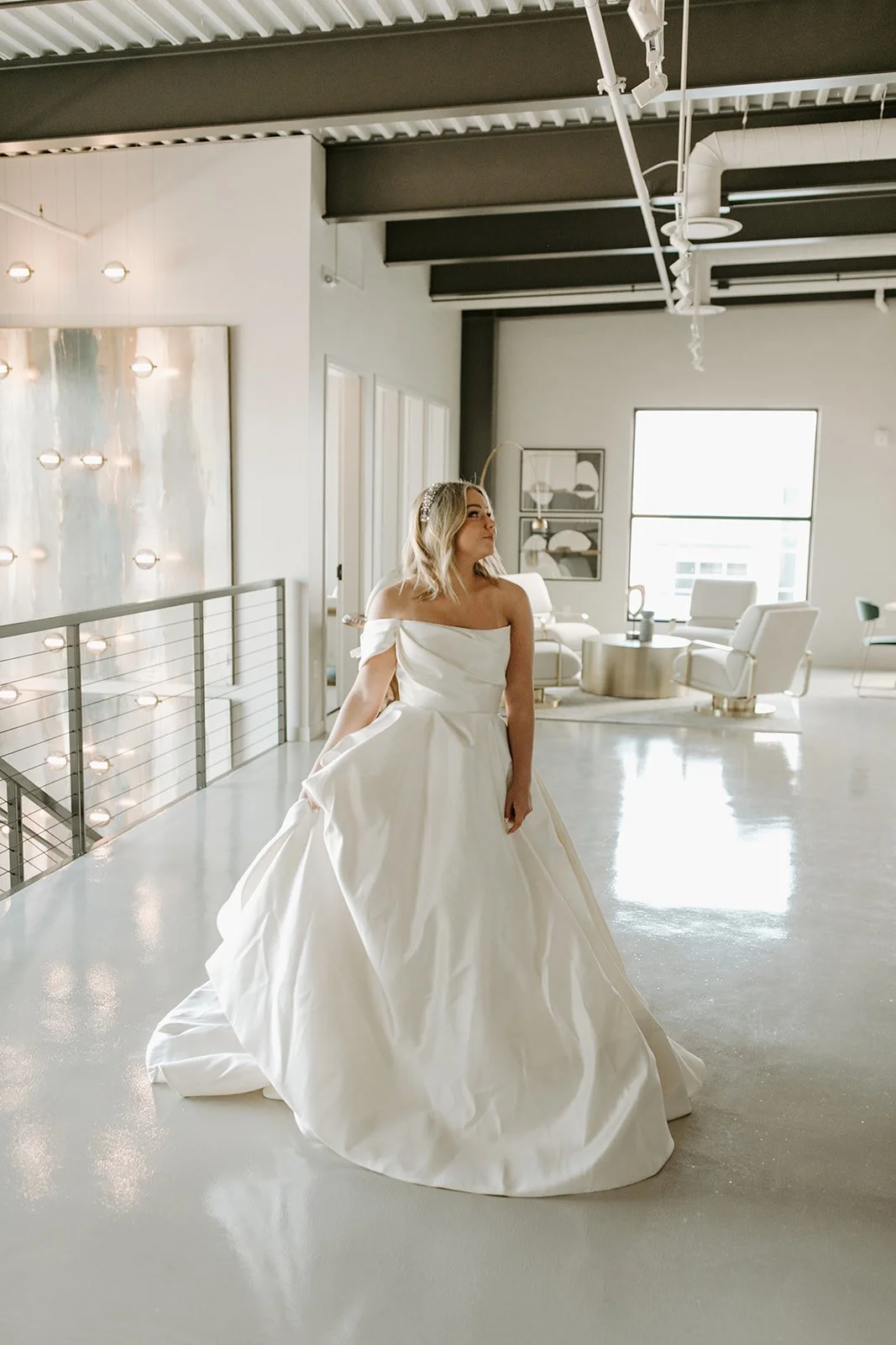 A woman in a white off-shoulder wedding dress standing in a modern, bright room with minimalist decor.