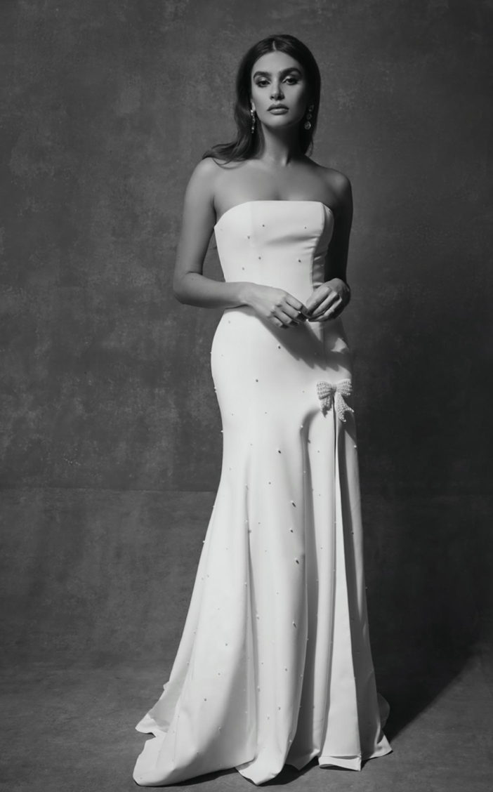 A woman wearing a strapless, floor-length white gown with a small bow and decorative details, standing against a textured gray background.