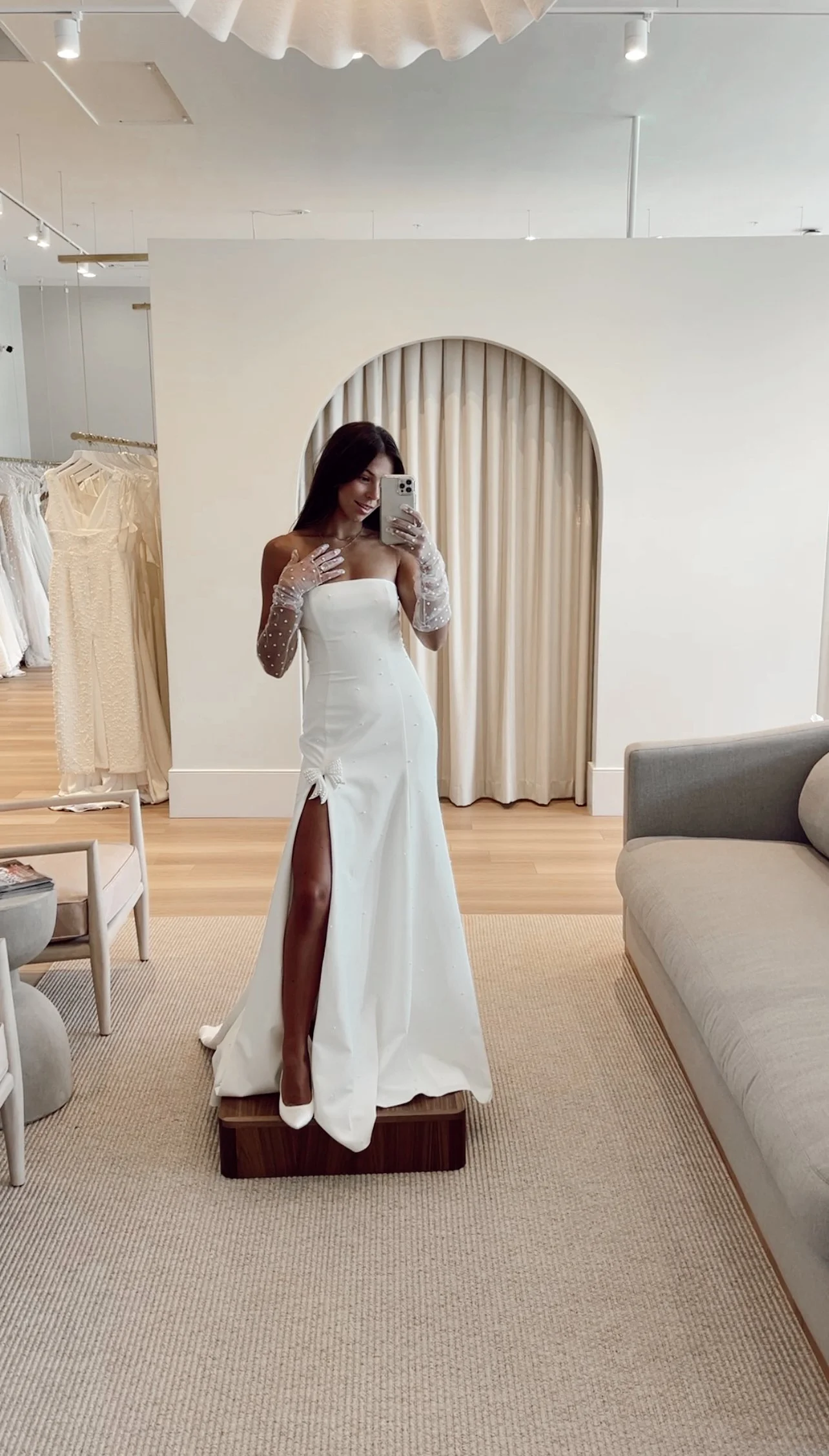 A woman taking a mirror selfie while trying on a white wedding dress with a high slit, lace gloves, and white heels in a boutique fitting room.
