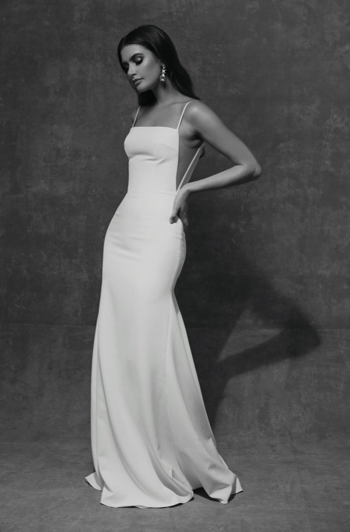 A woman in a white evening gown with spaghetti straps and cutout sides, standing against a textured dark background. She has long dark hair, prominent earrings, and is looking down with a serious expression.