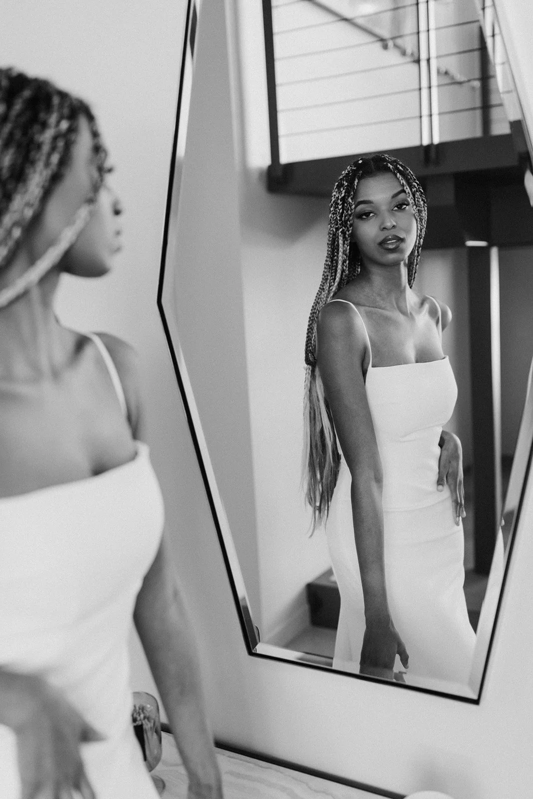 A woman with braided hair looking at her reflection in a mirror, wearing a light-colored strapless dress, black lipstick, in a modern interior with staircase railing in the background.