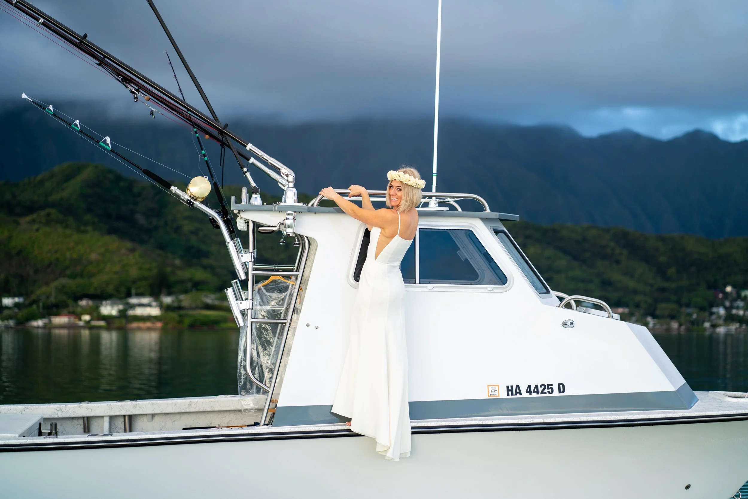 A woman in a white dress wearing a flower crown stands on a boat with fishing rods, smiling and holding onto the boat's structure, with mountains and a cloudy sky in the background.