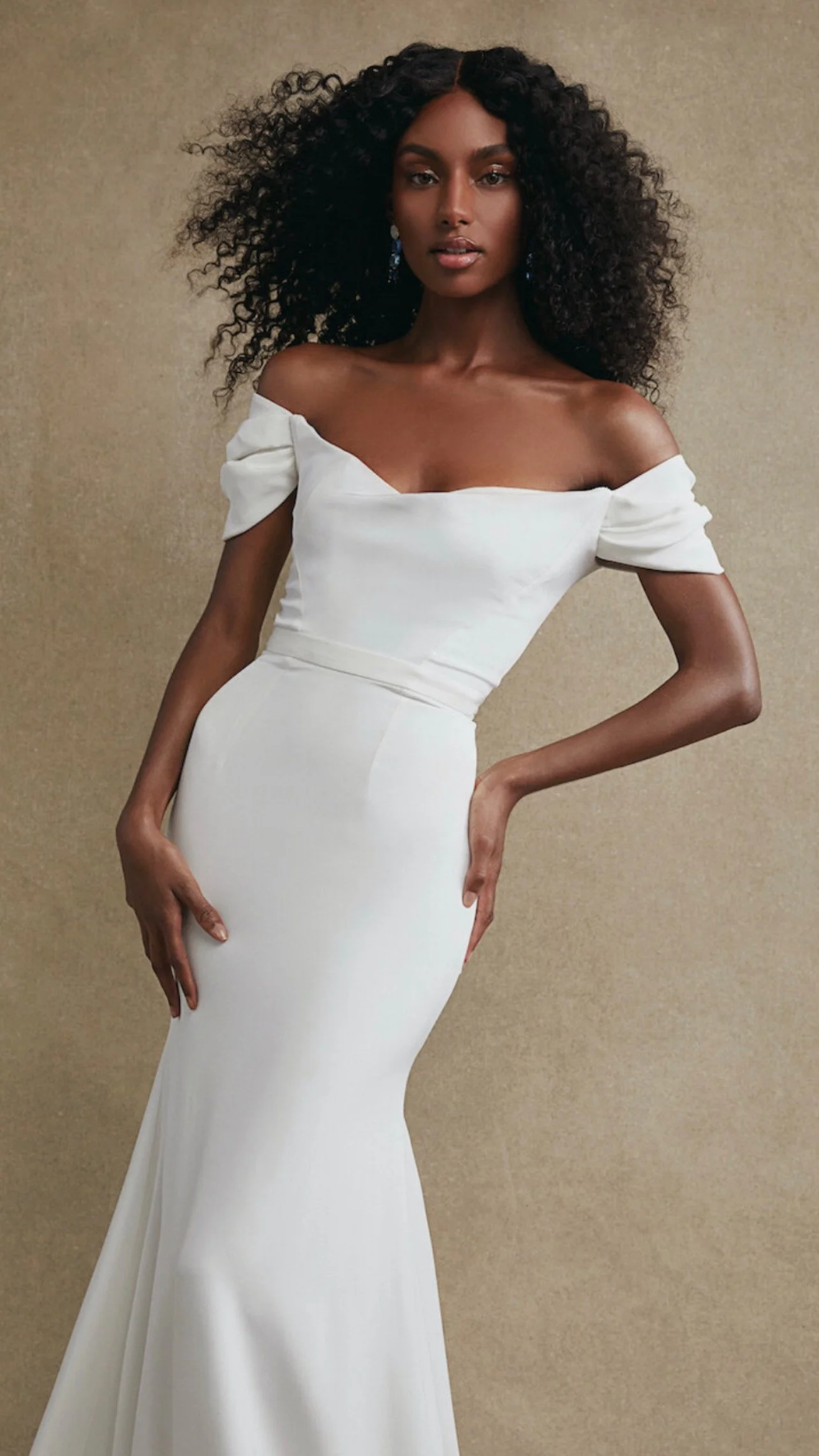 A woman with dark skin and voluminous curly hair wearing a white off-the-shoulder dress against a plain beige background.