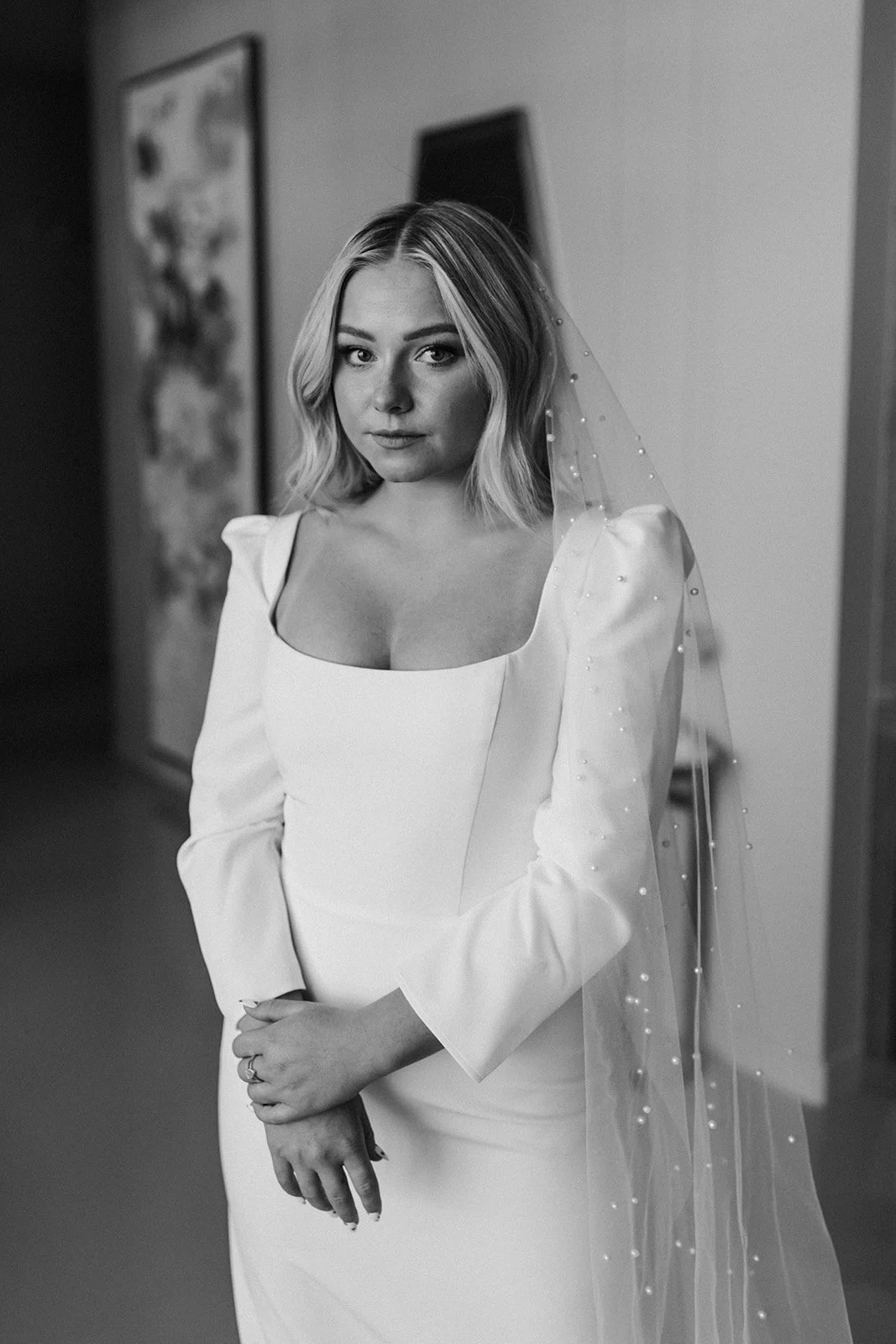 A woman in a wedding dress with puffed sleeves and a veil with pearls, standing indoors with artwork on the wall behind her.