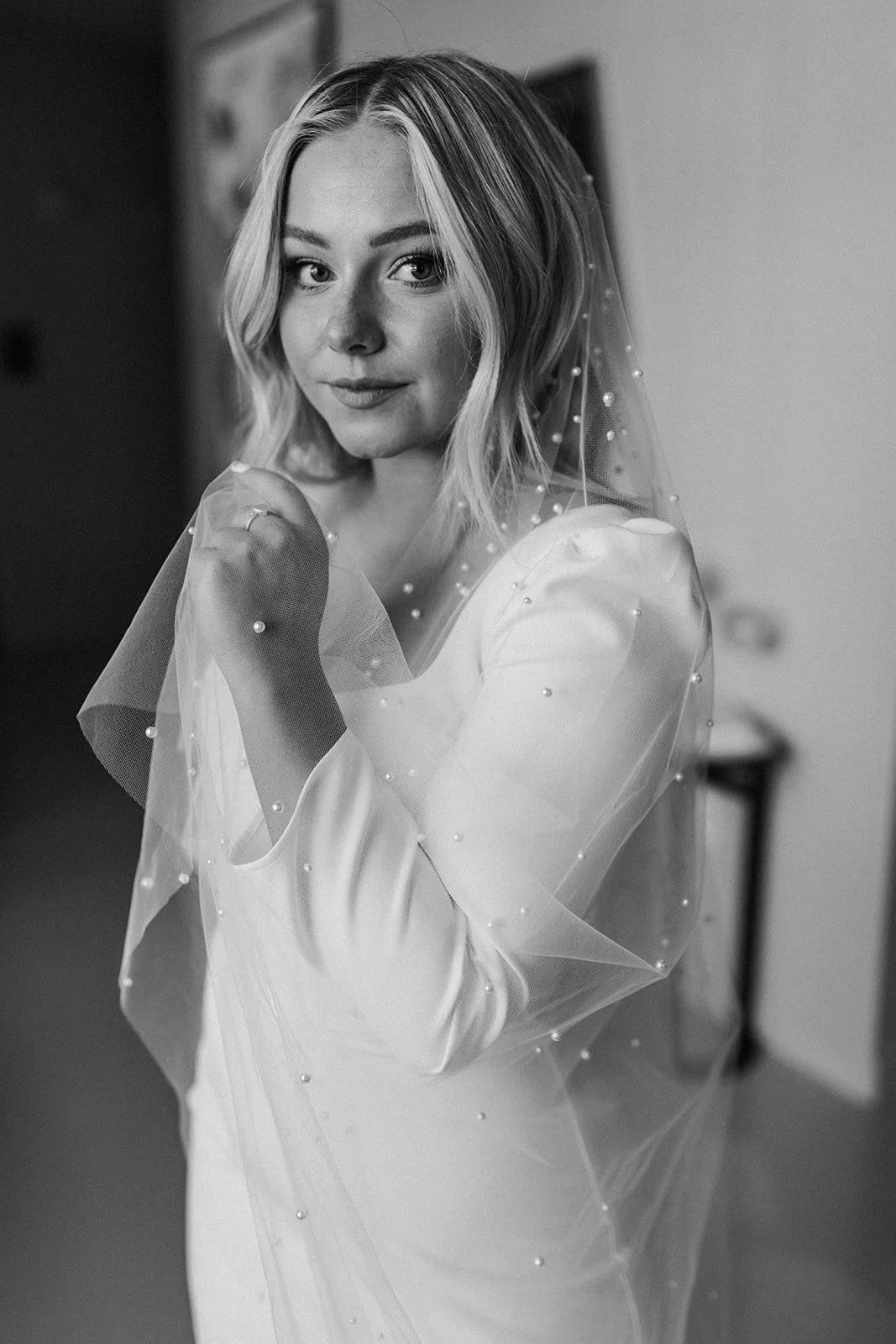 A woman with shoulder-length blonde hair wearing a wedding dress and veil with pearls, standing indoors and looking at the camera.