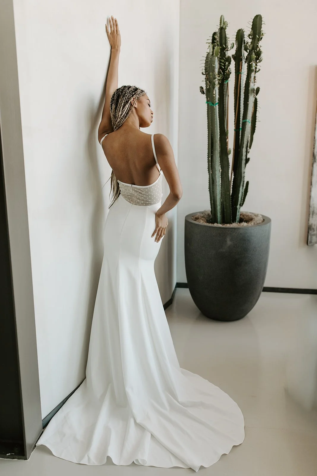 A woman in a white wedding dress with a long train standing next to a large potted cactus, leaning against a white wall, with her hair styled in braids.