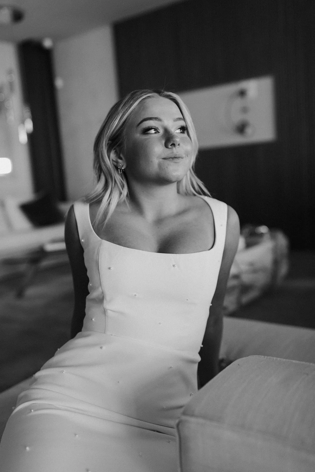 A woman in a sleeveless dress sitting on a couch in a modern room, looking to the side with a slight smile.