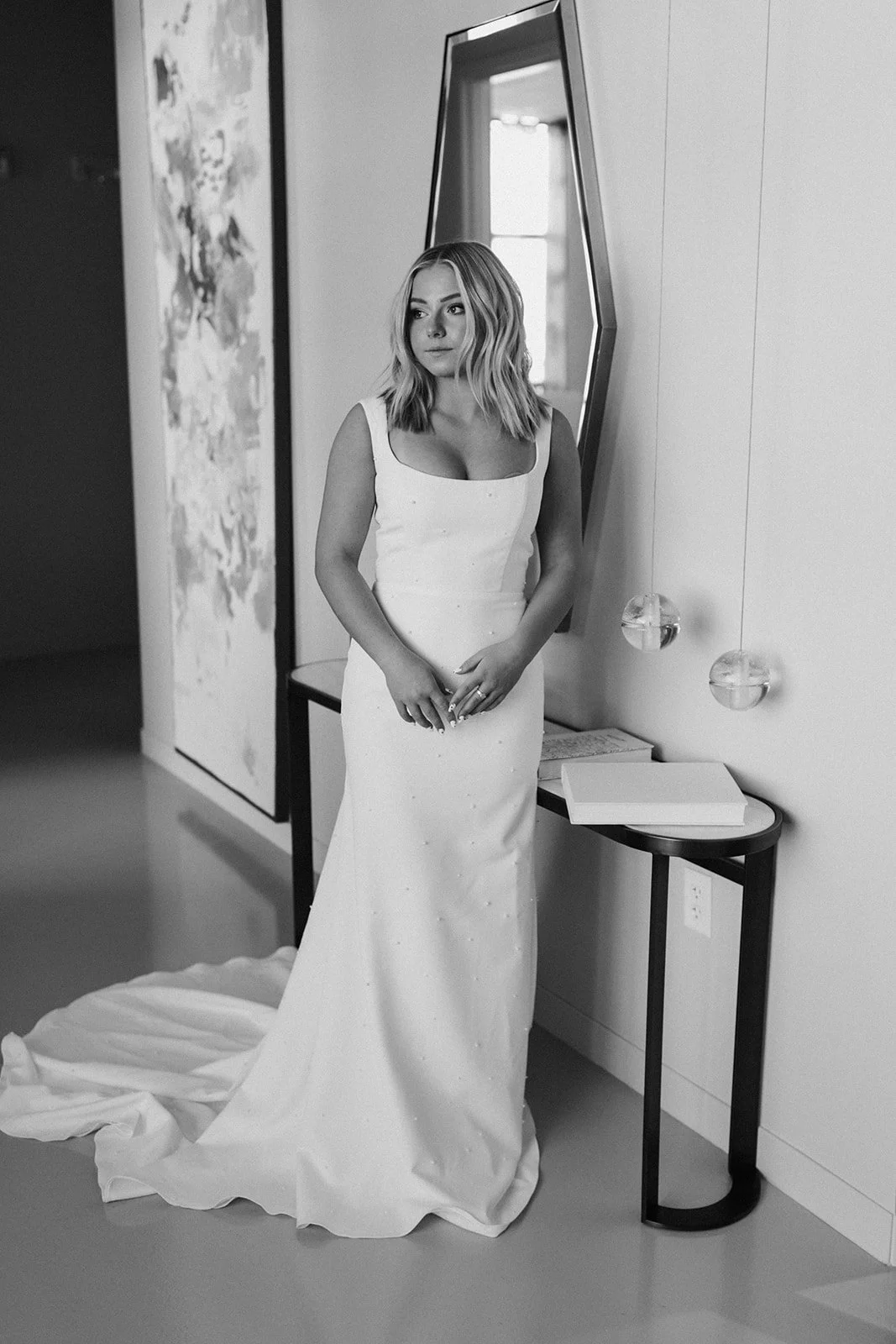 Black and white photo of a woman in a wedding dress standing in a modern interior near a mirror and a side table with books.