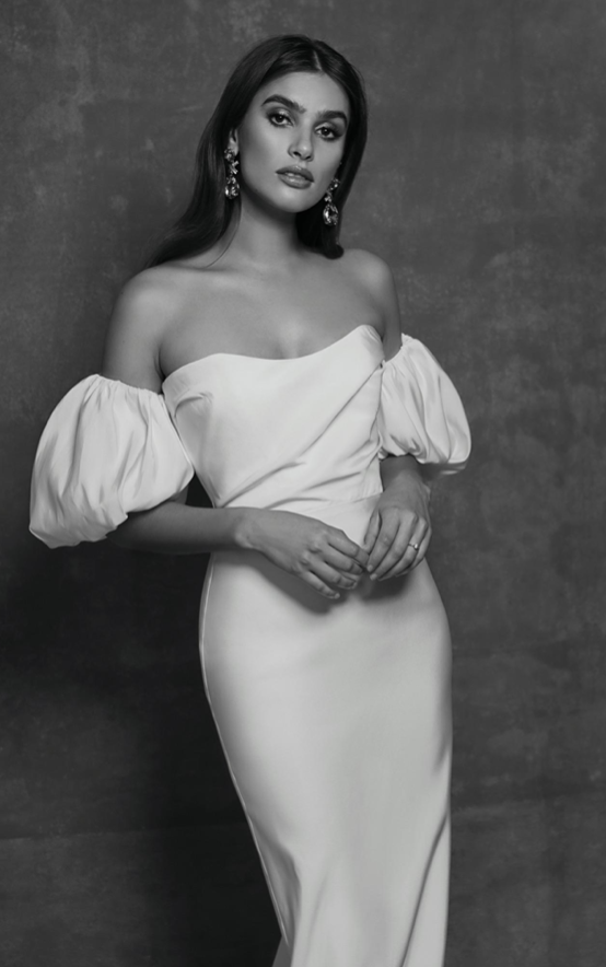 A woman in an elegant off-the-shoulder gown with puffy sleeves, wearing large earrings, posing against a textured background in black and white.