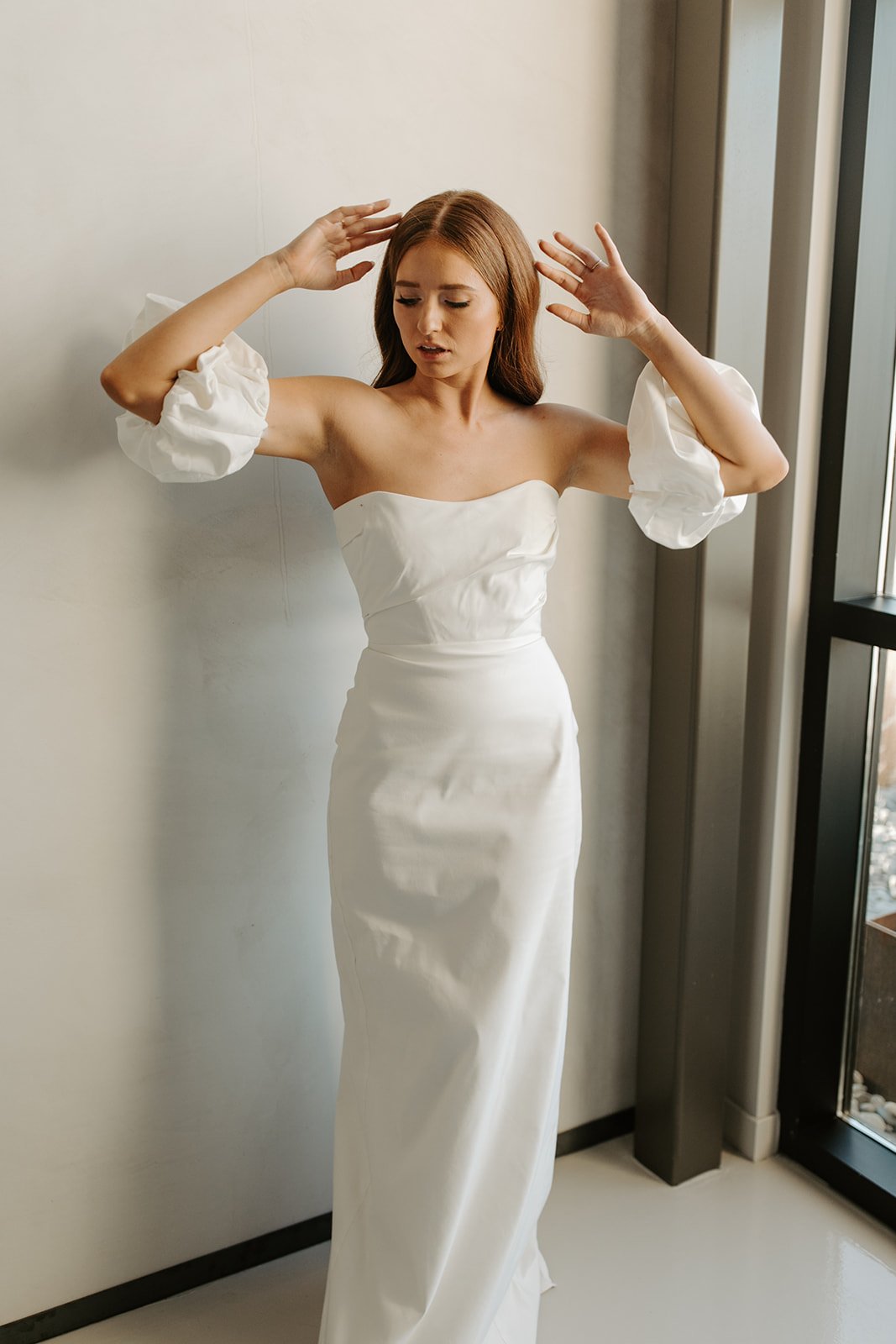 A woman with red hair wearing a strapless white dress with puffed sleeves standing indoors near a window, with her arms raised and hands near her head.