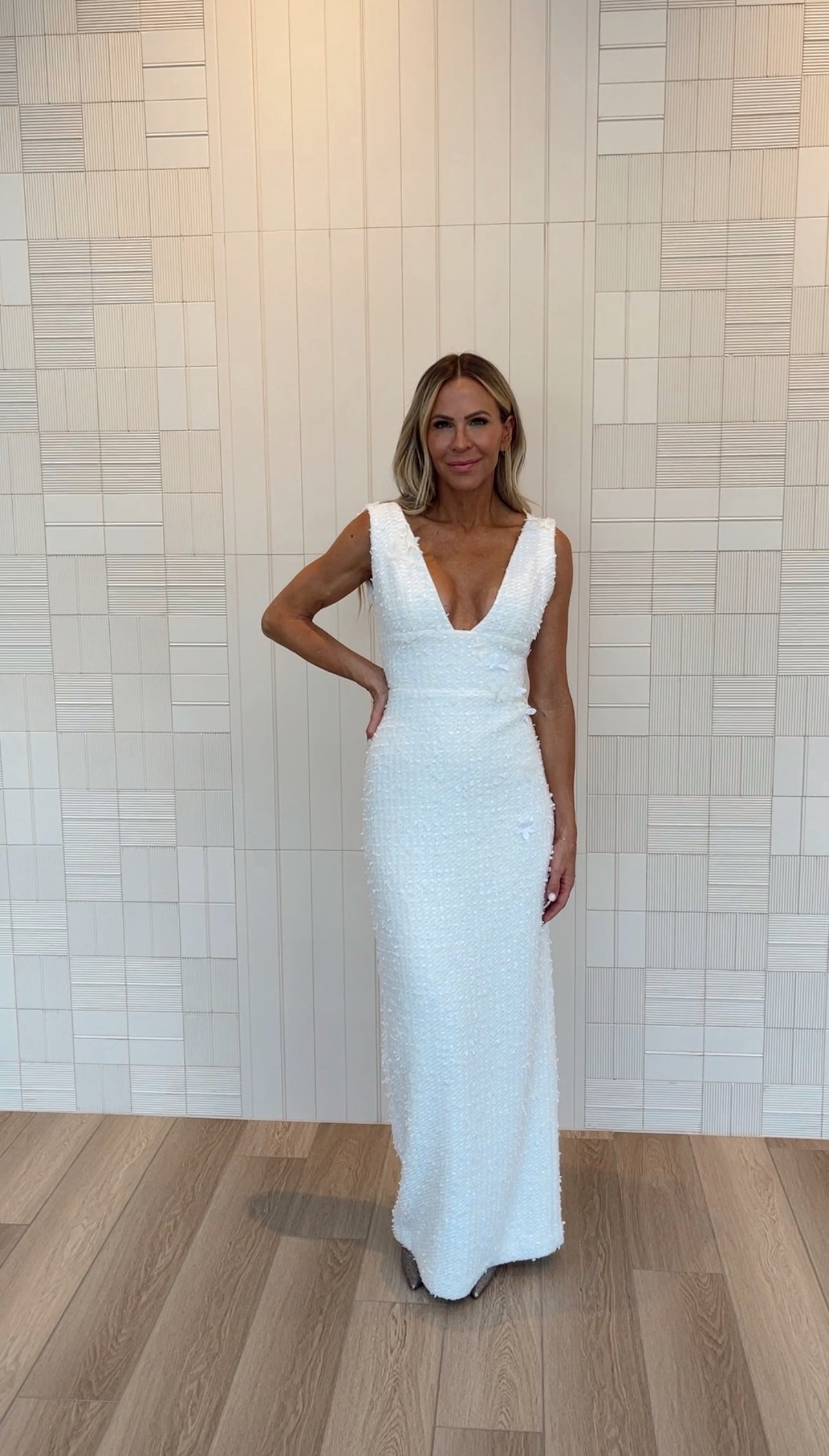 Woman in a long white dress standing against a beige tiled wall.