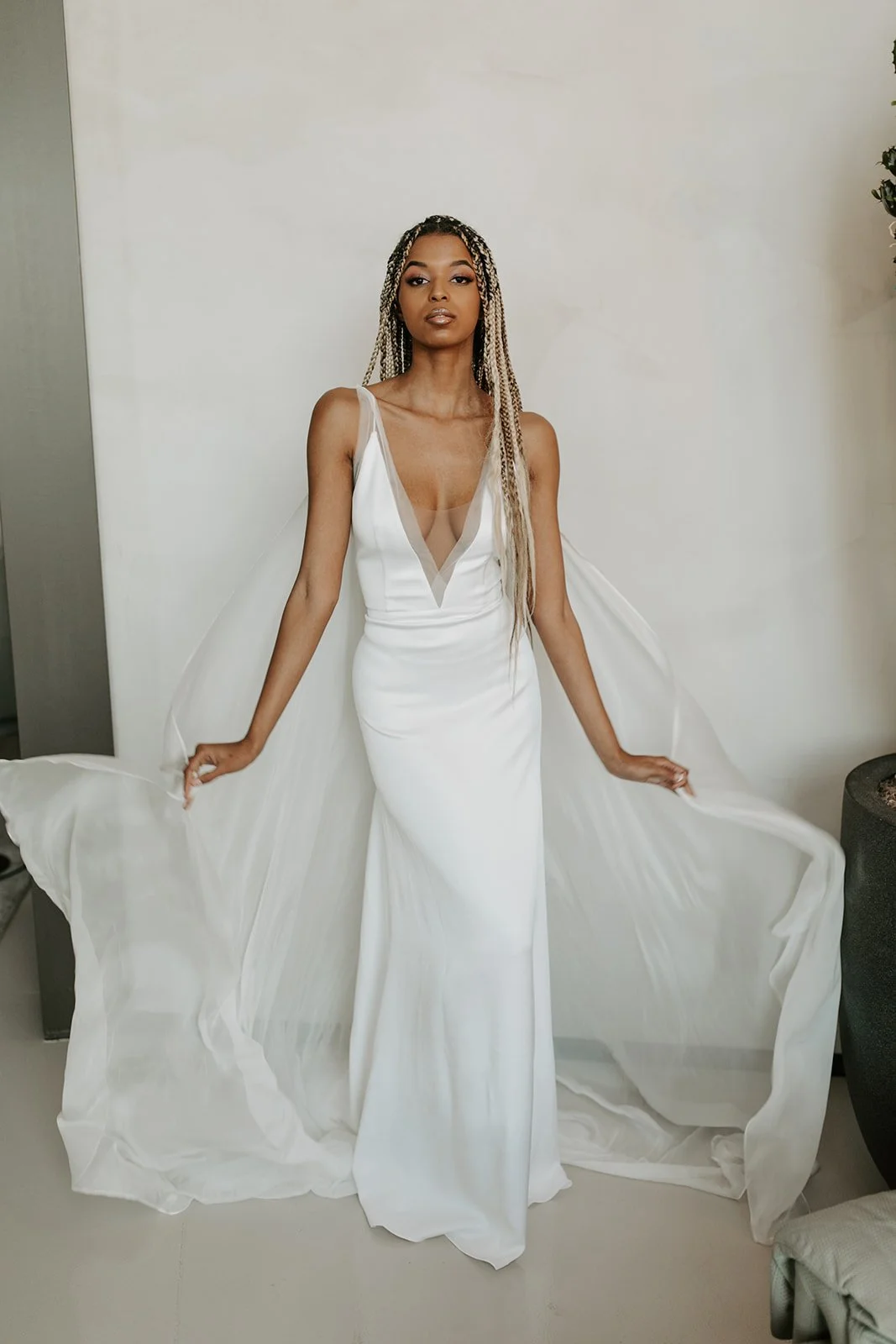 A woman in a white gown standing against a plain wall, holding the sides of her dress.