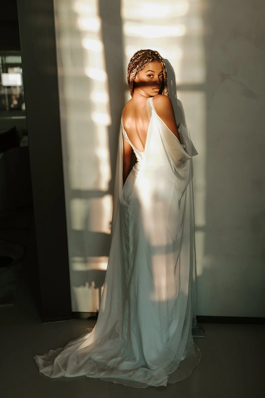 A woman in a white dress stands near a wall with sunlight casting shadows on her.