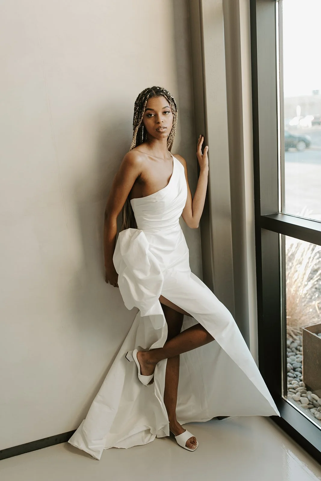 A woman in a white dress with a high slit, standing by a large window with natural light, in a minimalistic interior setting.