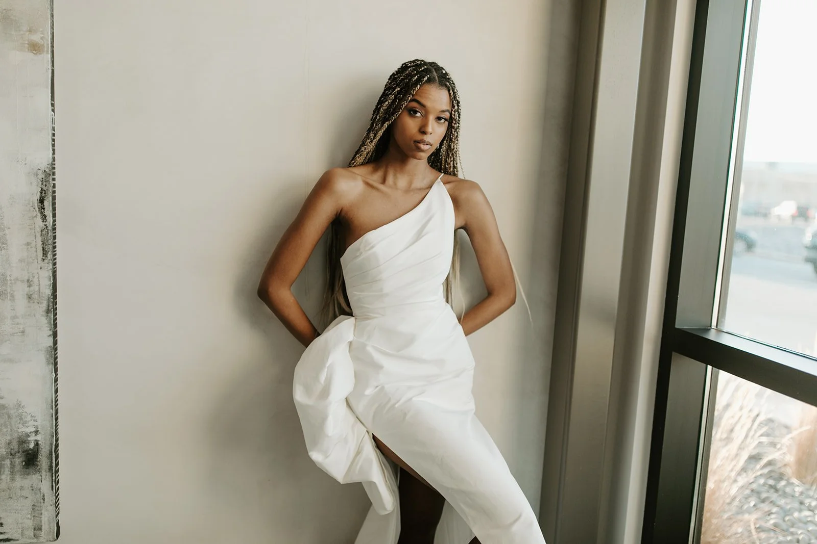 A woman with long braided hair leaning against a wall near a window, wearing a white dress.