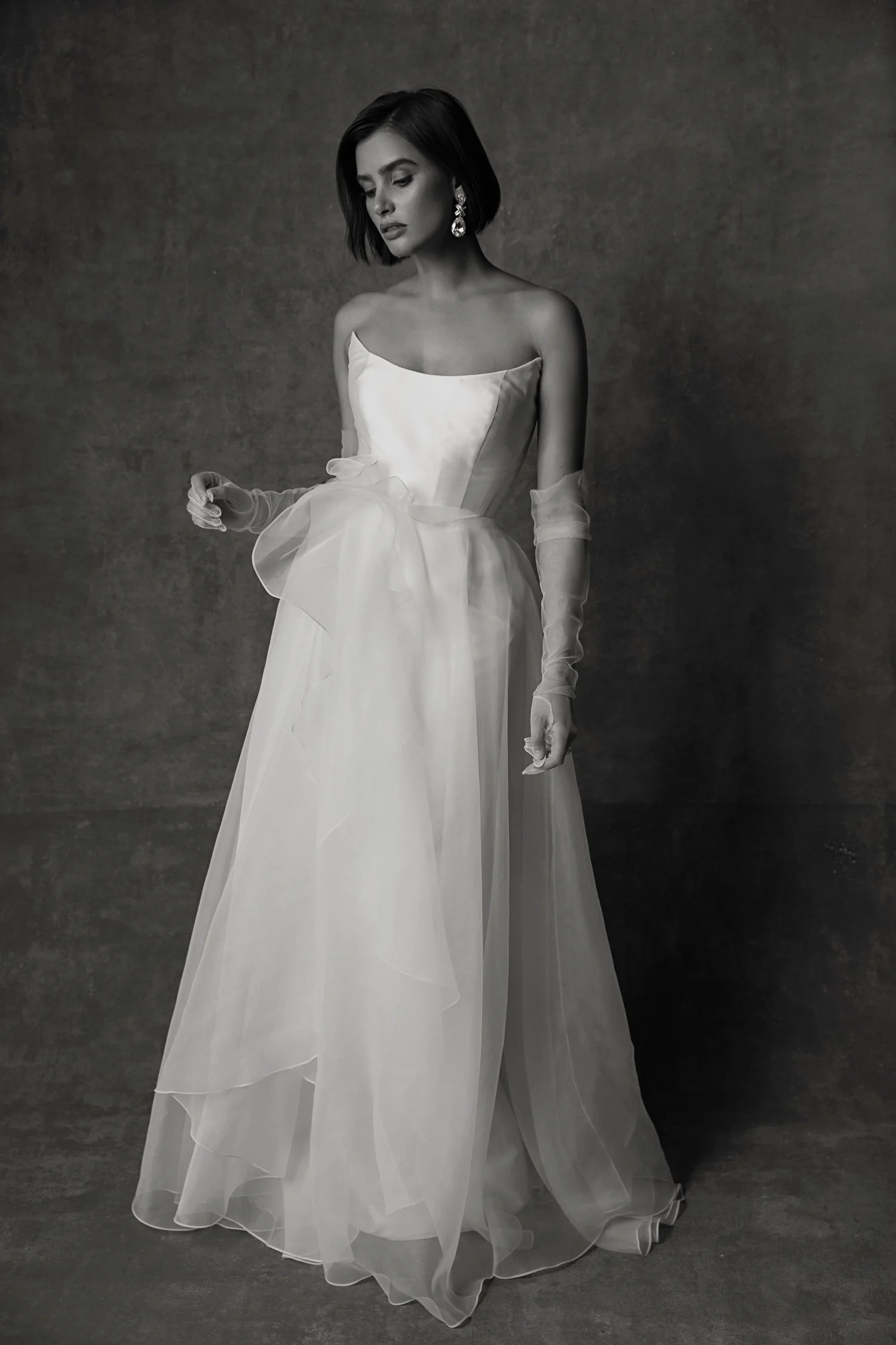 A woman wearing a strapless wedding gown with a bow at the waist, long sheer gloves, and large earrings stands against a plain dark background in a black and white photo.