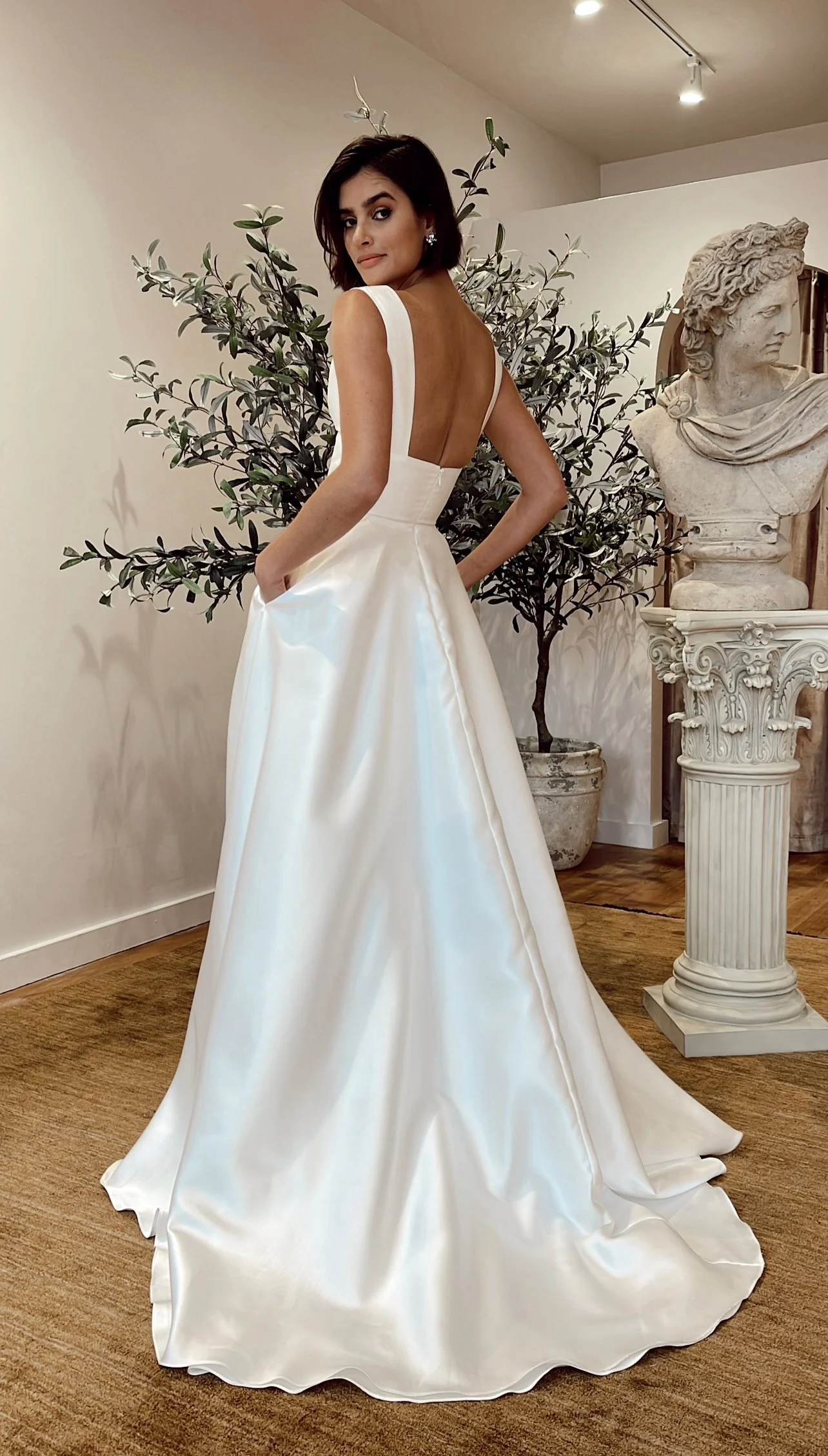 A woman in a white satin gown with a fitted bodice and a full skirt, standing indoors with a classical sculpture and greenery in the background.