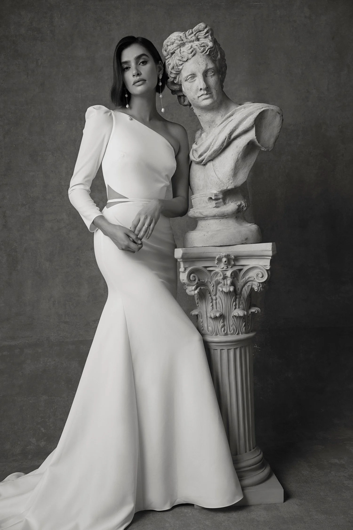 A woman in a white wedding gown standing next to a classical stone bust sculpture on a decorative pedestal.