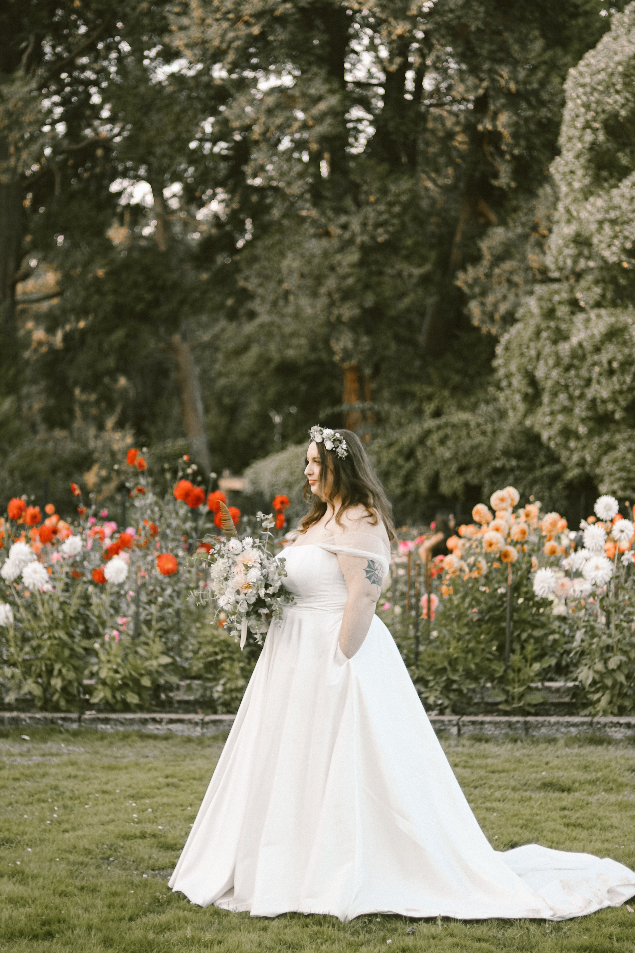 A bride in a white wedding dress with a floral crown, holding a bouquet, standing in a garden with colorful flowers and greenery.