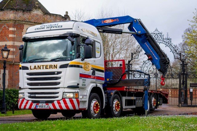 A large white and blue crane truck with red and white striped bumper, parked on a grassy area near a brick building and decorative iron gate.