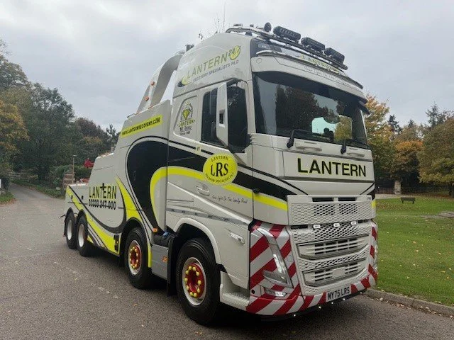 A large truck with a white body, black and yellow stripes, and red and white striped front bumper, parked on a street with trees and grass in the background. The truck has the words "LANTERN" and "LRS" on it, with additional text and logos.