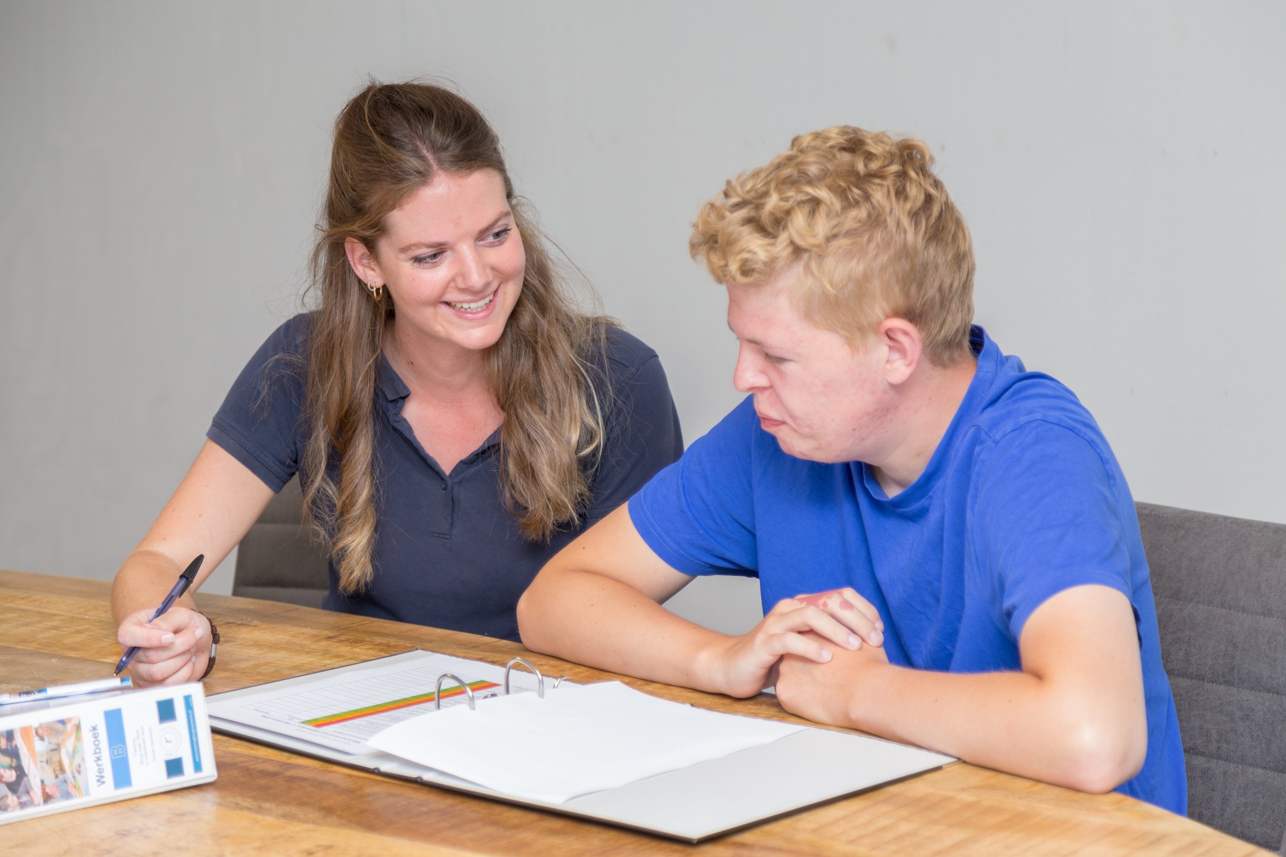 Een vrouw met lang bruin haar en een man met kort, krullend blond haar zitten samen aan een tafel. De vrouw glimlacht terwijl ze met een pen in haar hand een document vasthoudt, en de man kijkt aandachtig naar het document. Op de tafel liggen papieren en een ringband, en er staat een informatiekaart op de voorgrond.