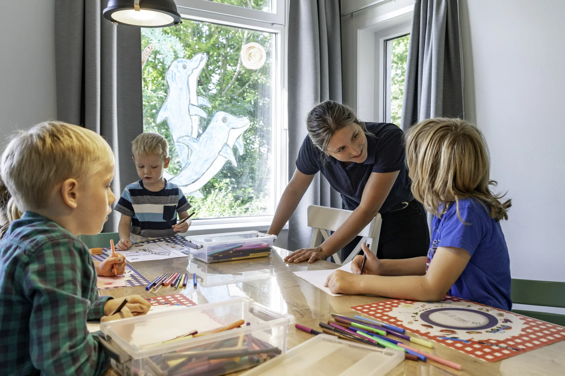 Vrouw geeft kinderen tekenles in een lichte kamer met grote ramen en een decoratief houten zeehondenkunstwerk buiten.