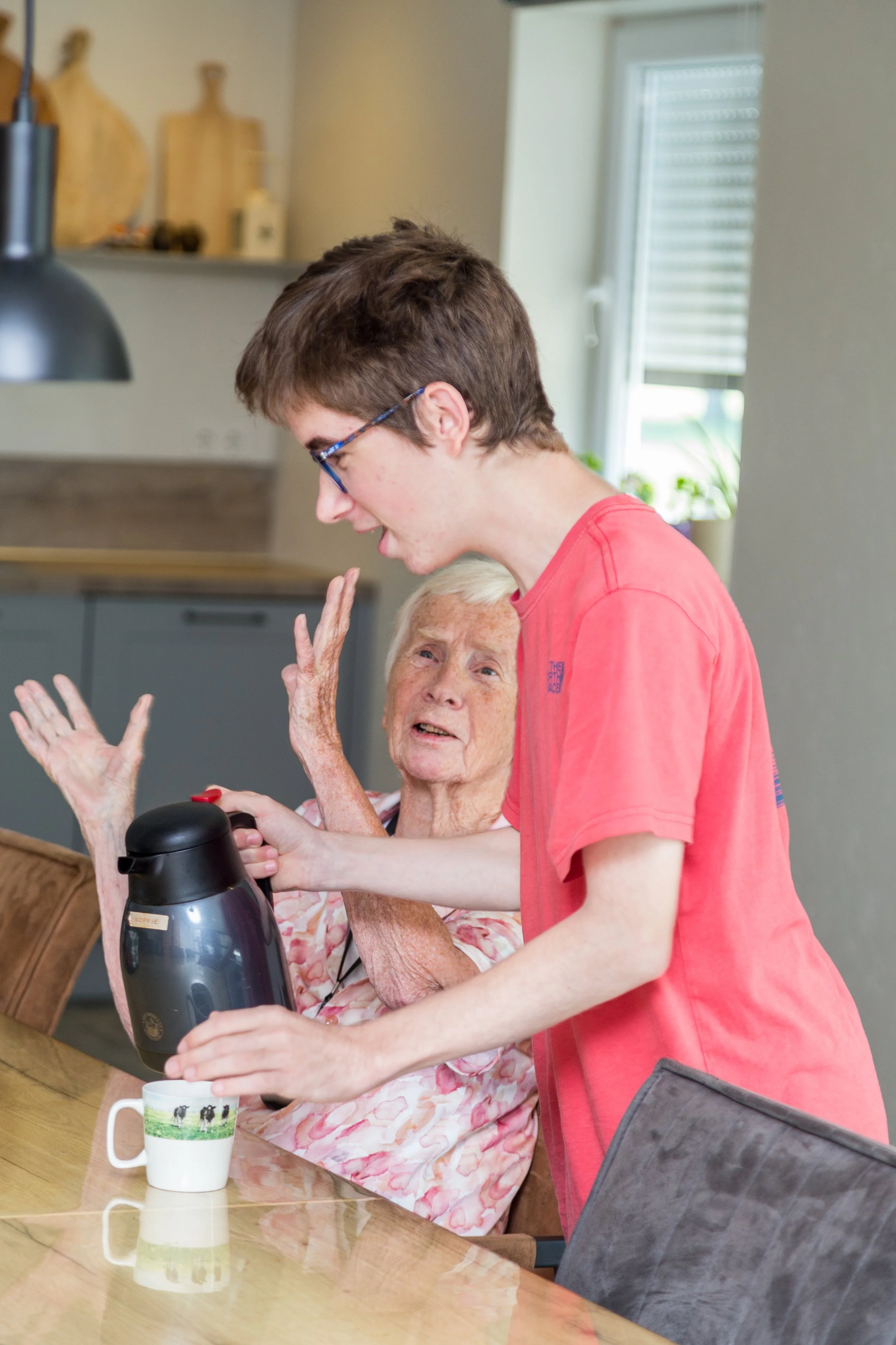 Een jonge man helpt een oudere vrouw met het inschenken van koffie aan de keukentafel. De vrouw zit en de man staat naast haar, beiden lijken blij en betrokken.