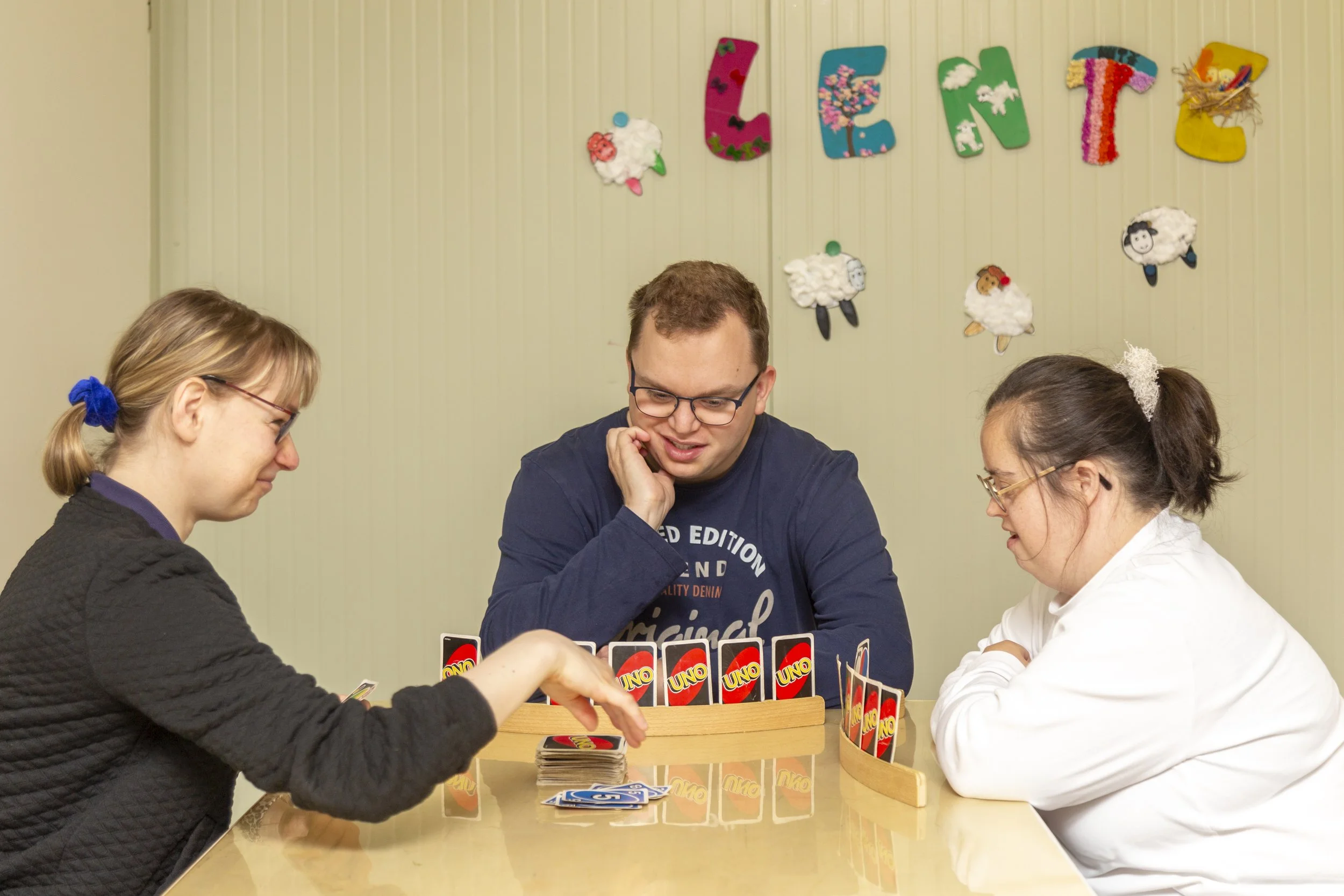 Vier mensen spelen een potje UNO kaartspel aan een tafel. Op de achtergrond hangen decoraties in de vorm van letters en schapen van katoen op de muur.