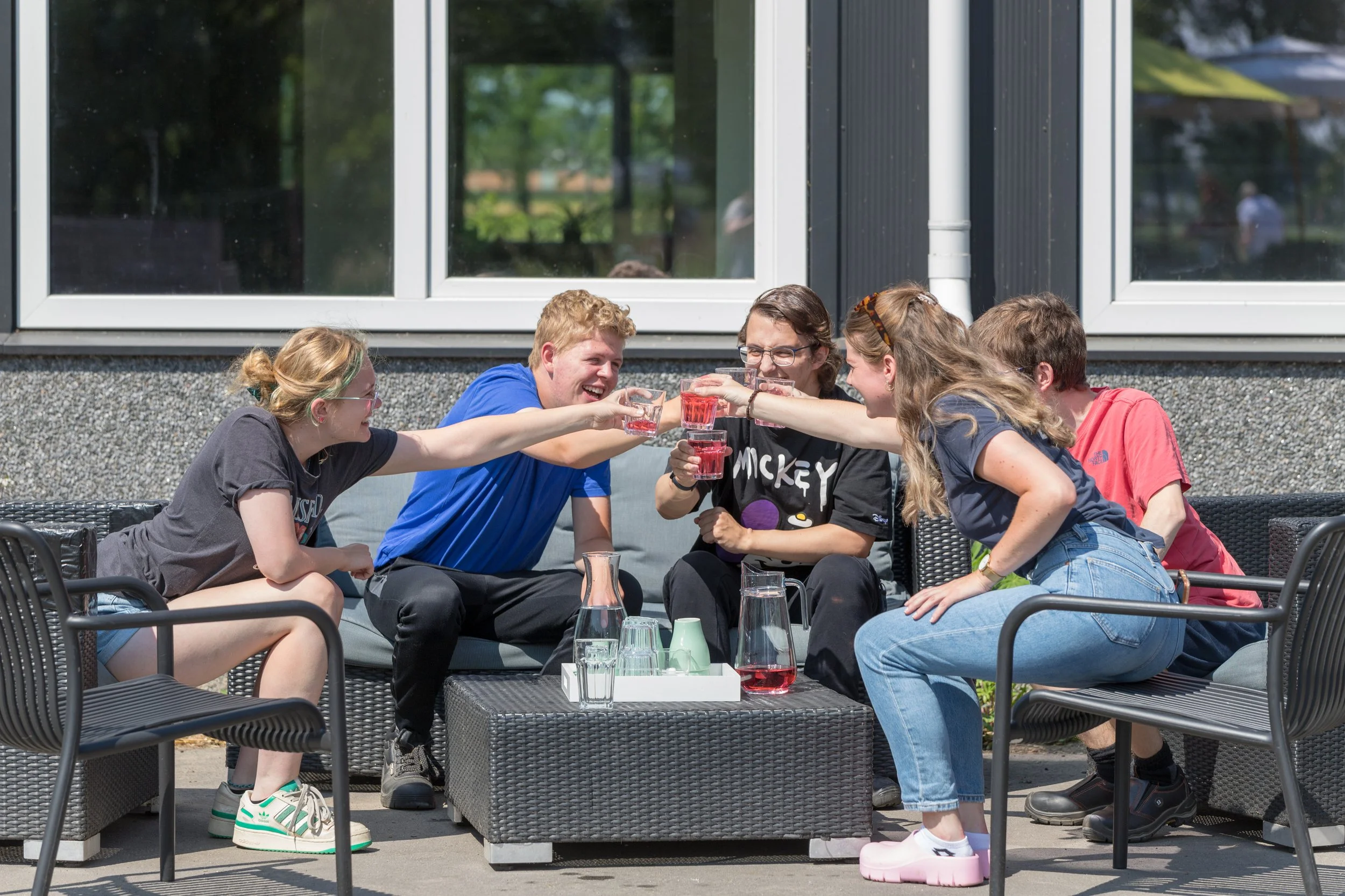 Vijf jonge mensen zitten buiten op een terras en heffen hun glazen met een rood drankje naar elkaar op in een toast, terwijl ze lachen en plezier hebben.