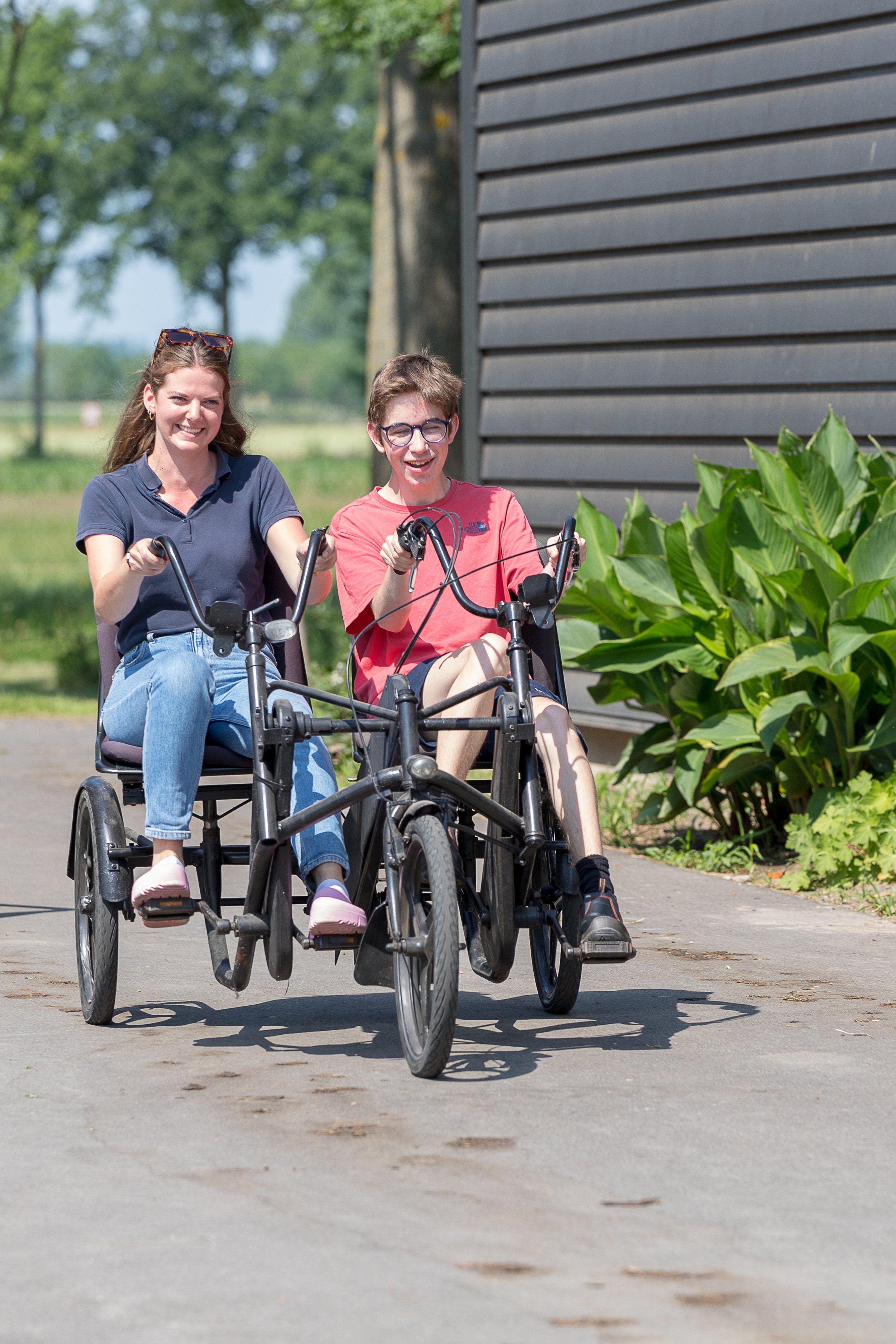 Twee mensen rijden in een aangepaste driewieler op een pad buiten met groene planten en bomen op de achtergrond.