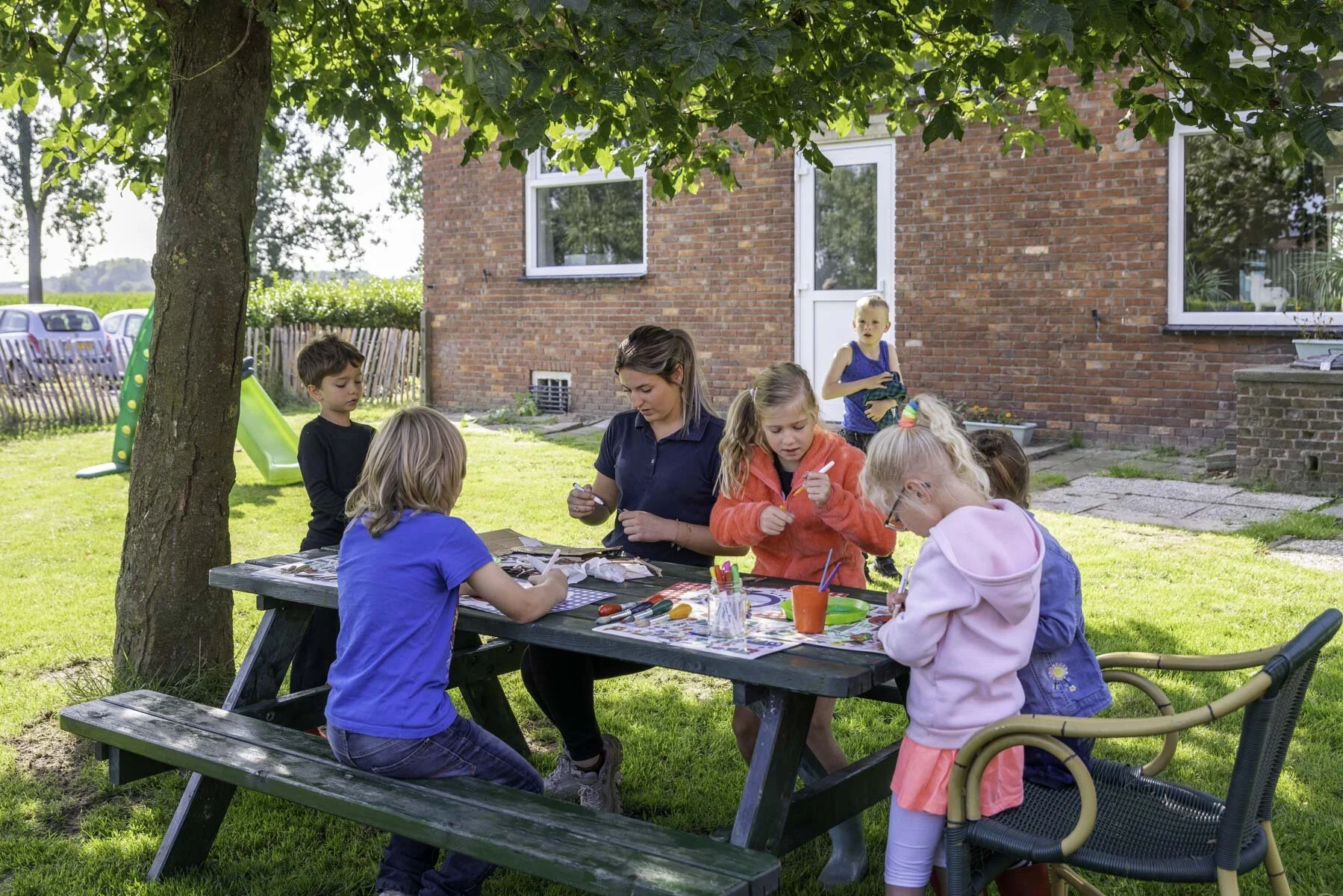 Kinderen en een begeleider knutselen buiten aan een picknicktafel onder een boom, met een huis op de achtergrond.