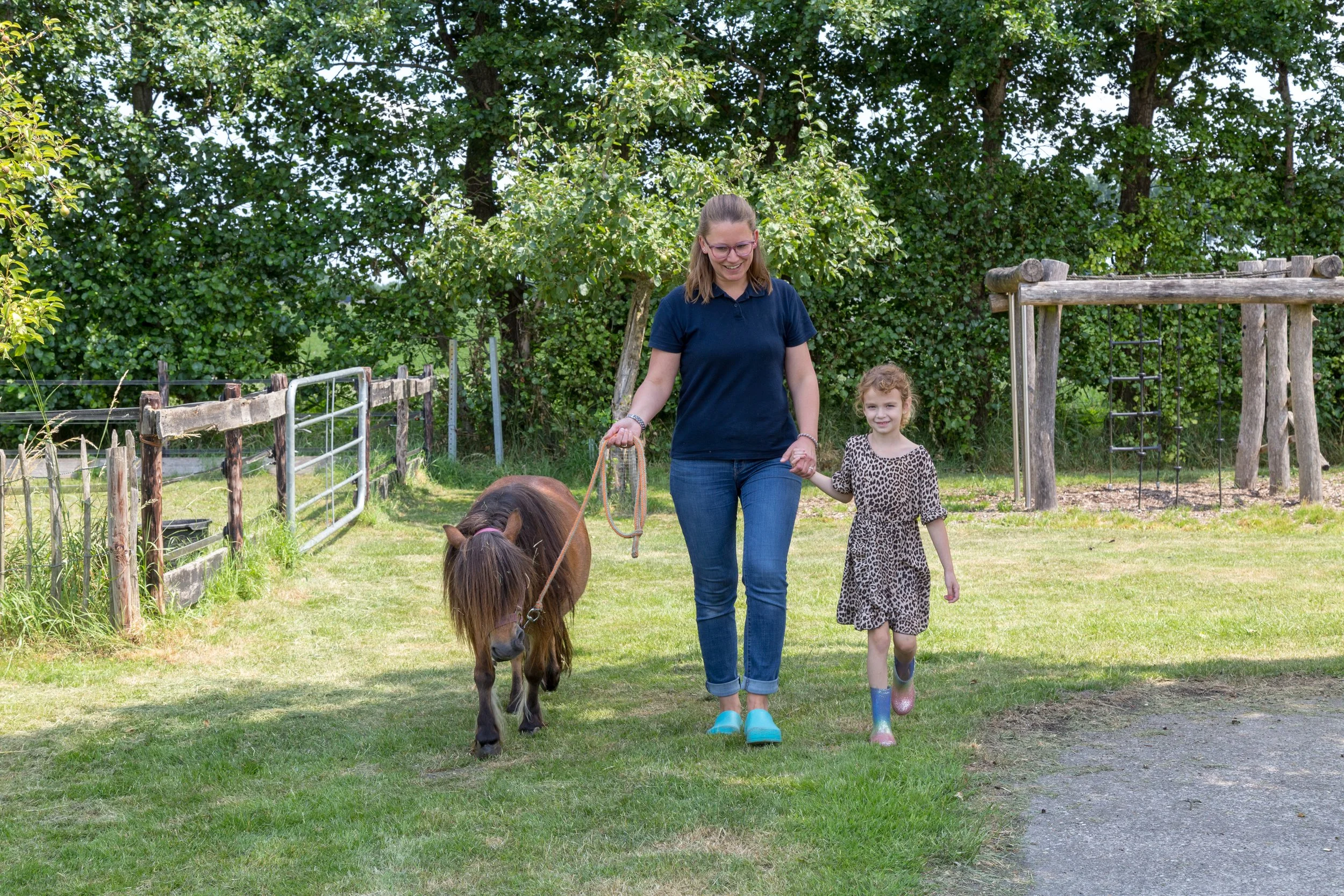Vrouw en kind wandelen op een grasveld in een park, met een paardje naast hen.