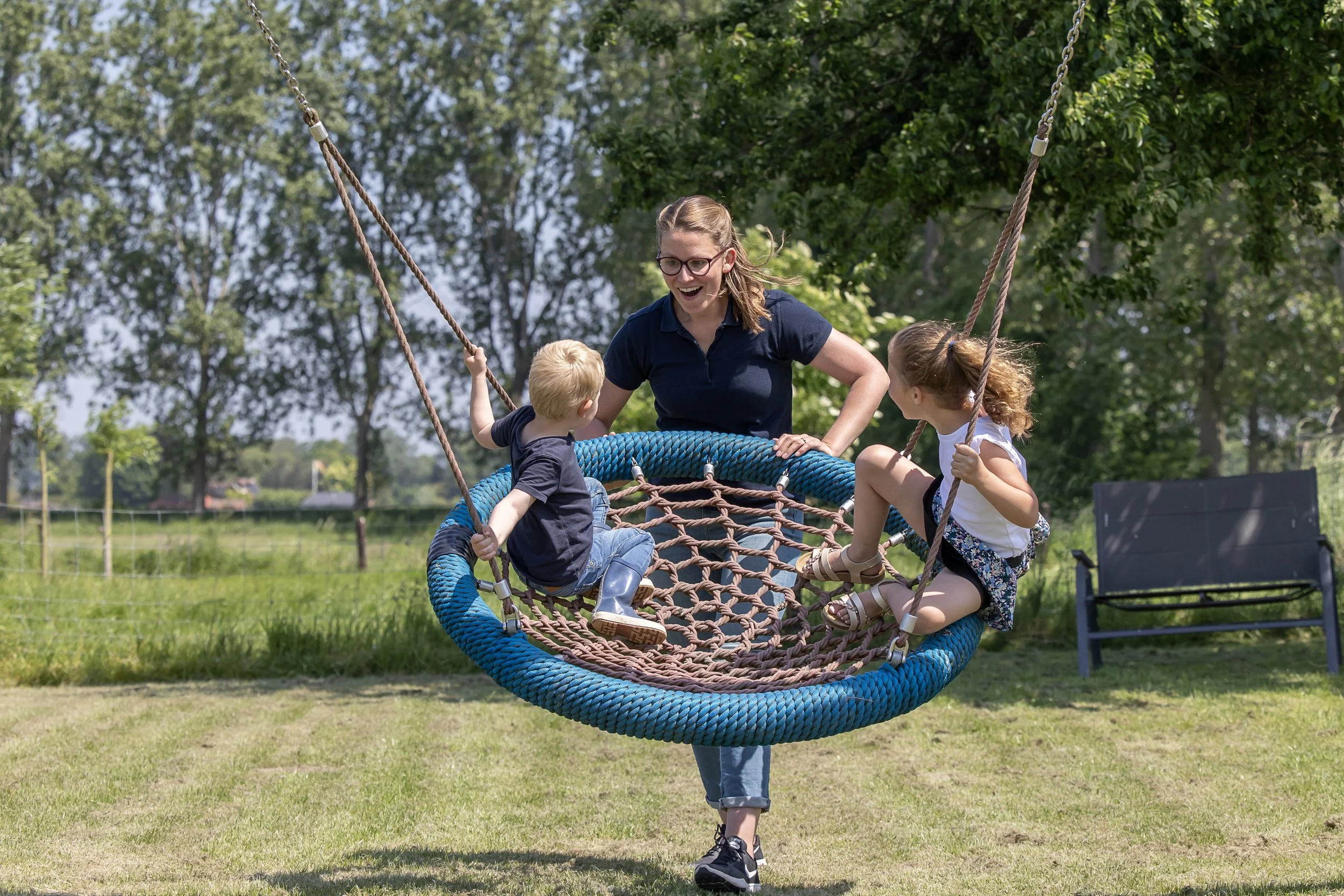 Vrouw met twee kinderen die op een schommel hangen in een park, omringd door bomen en gras, glimlachend en genietend van het buiten zijn.