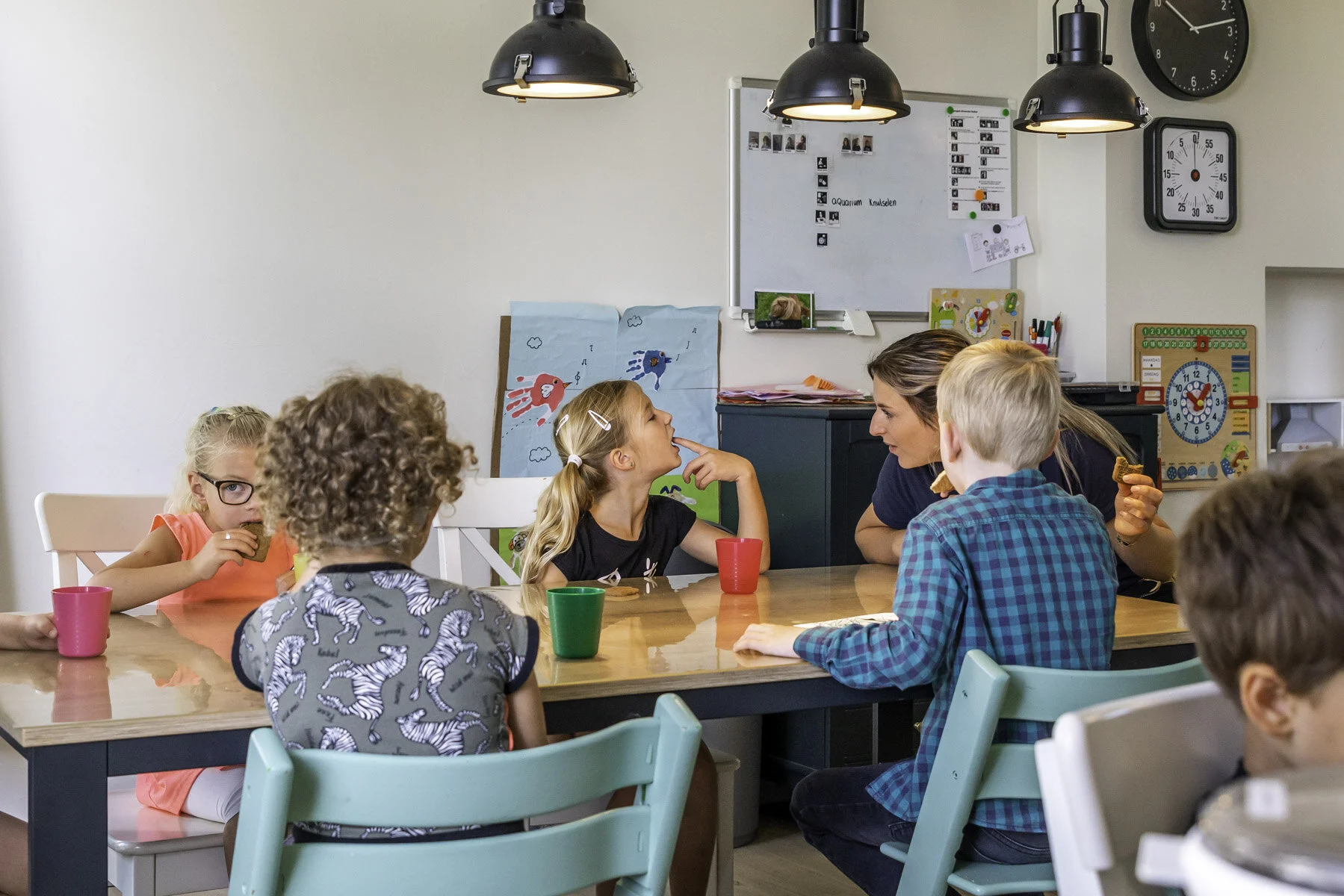 Kinderen en een volwassene zitten aan een tafel in een klaslokaal, terwijl een vrouw met lang haar met een kind praat. Er hangen kindertekeningen en educatieve materialen aan de muur, en er zijn koplampen en een klok aan de muur.