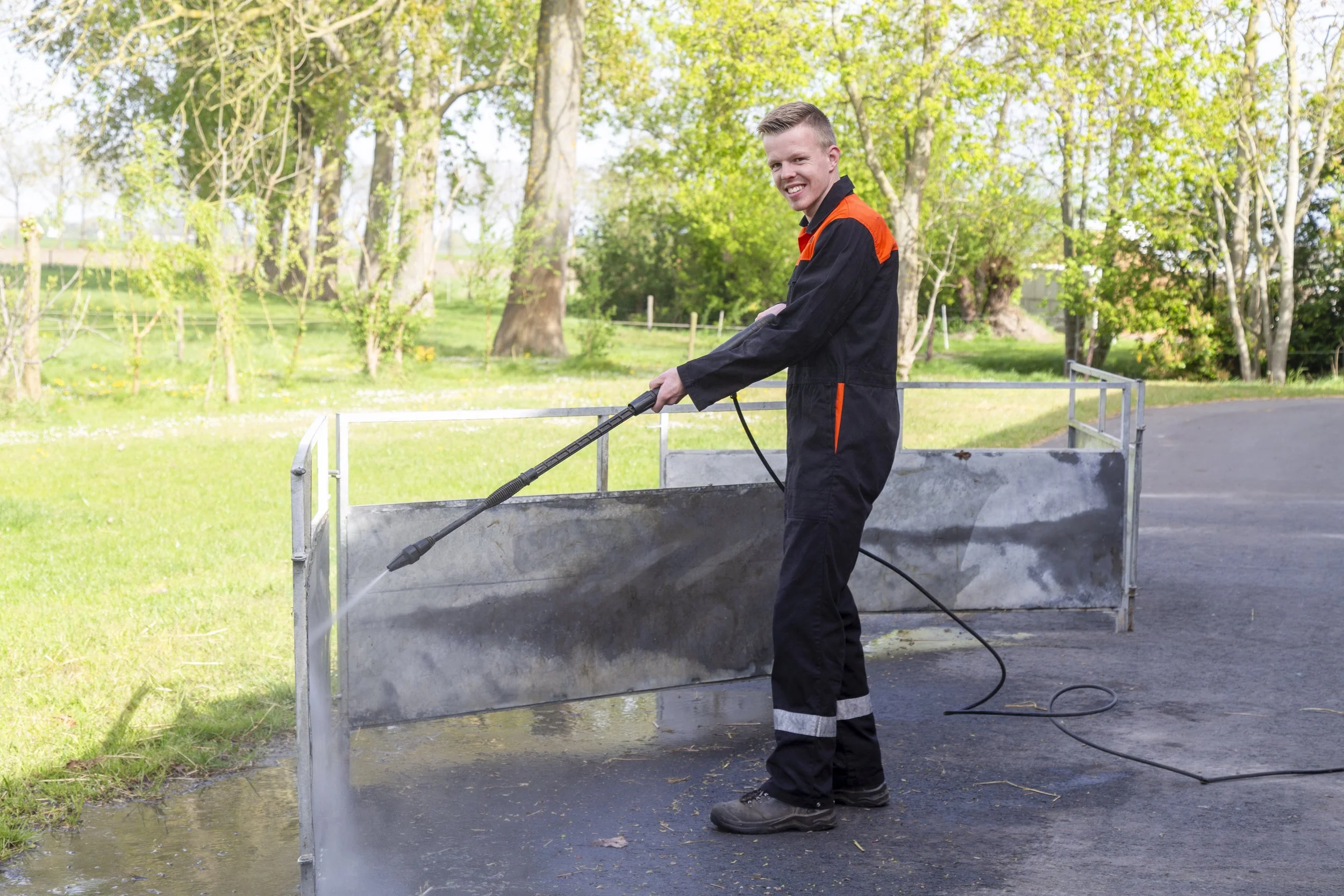 Man in werkoutfit met een hogedrukreiniger schoonmaakt op straat in een parkachtige omgeving met bomen en groen.