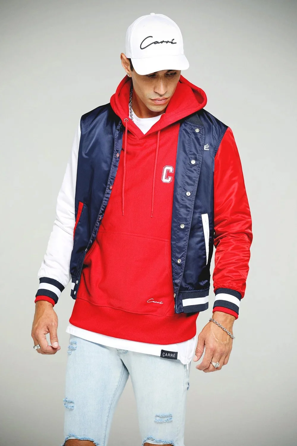 A young man wearing a white cap with "Carré" written on it, a red hoodie with a white "C" on it, a navy blue and red varsity jacket, and light blue ripped jeans.
