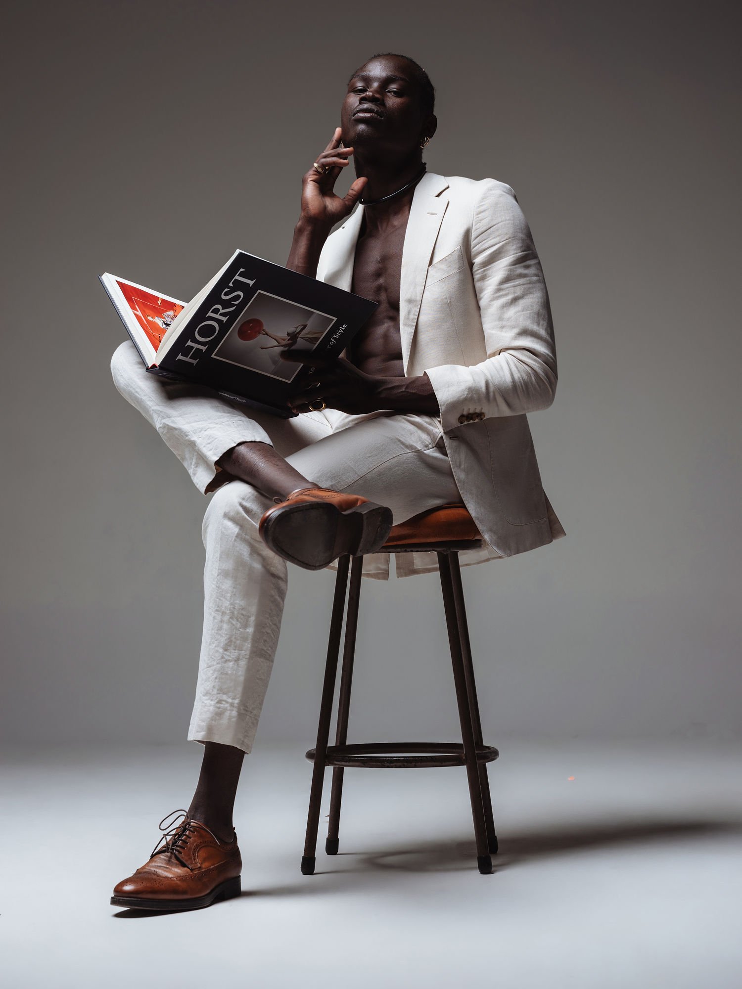 A man in a white suit is sitting on a stool, holding an open magazine titled "HORST". He is sitting with one leg crossed over the other, wearing brown dress shoes, and has a confident expression while touching his face with one hand.