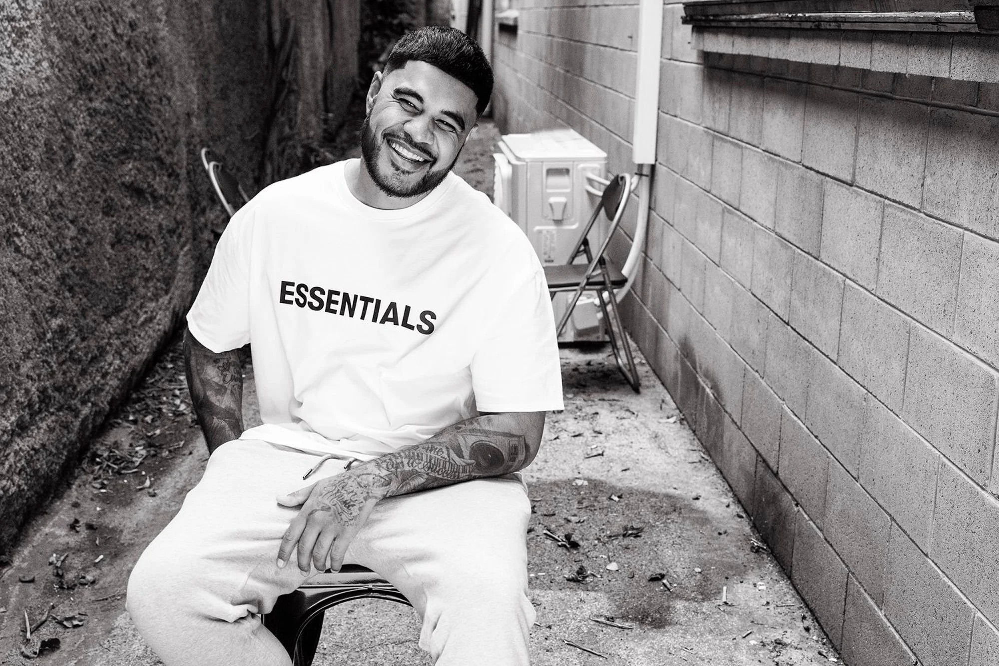 A smiling man with tattoos on his arms, wearing a white t-shirt with the word 'ESSENTIALS', sitting on a stool in an outdoor alley with a brick wall on one side and a textured wall on the other, with chairs and an outdoor air conditioning unit in the