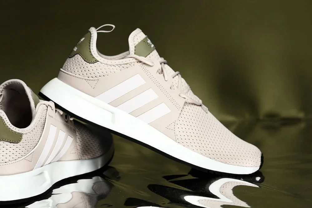 Beige athletic sneakers with white accents and a black sole, placed on a reflective surface.