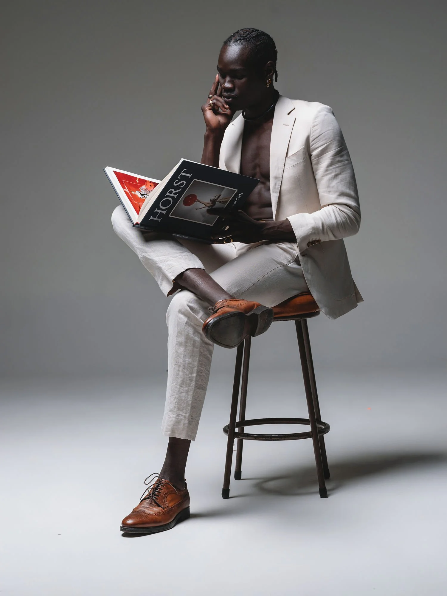 A person with dark skin, wearing a white suit with the jacket open, sits on a wooden stool against a plain gray background. They are reading a magazine titled "HORST." They are barefoot, with one foot wearing a brown dress shoe, and the other with no