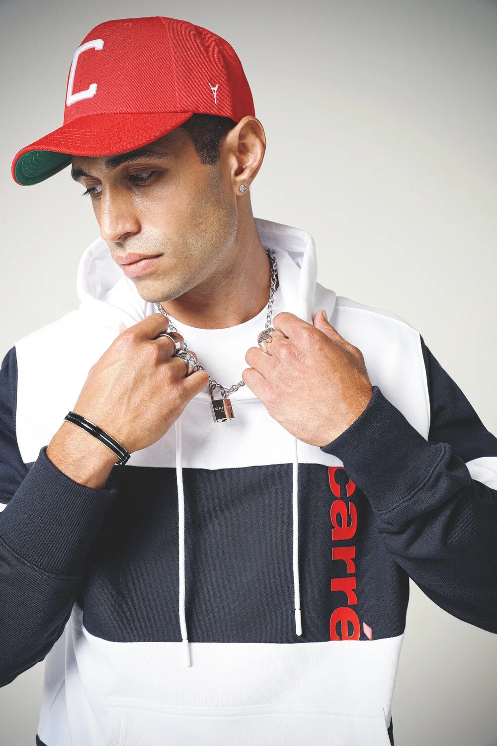 A young man with light brown skin, short dark hair, wearing a red baseball cap, a white and black hoodie with the word 'Supreme' on it, and accessories including rings, bracelets, and a chain with a lock pendant.