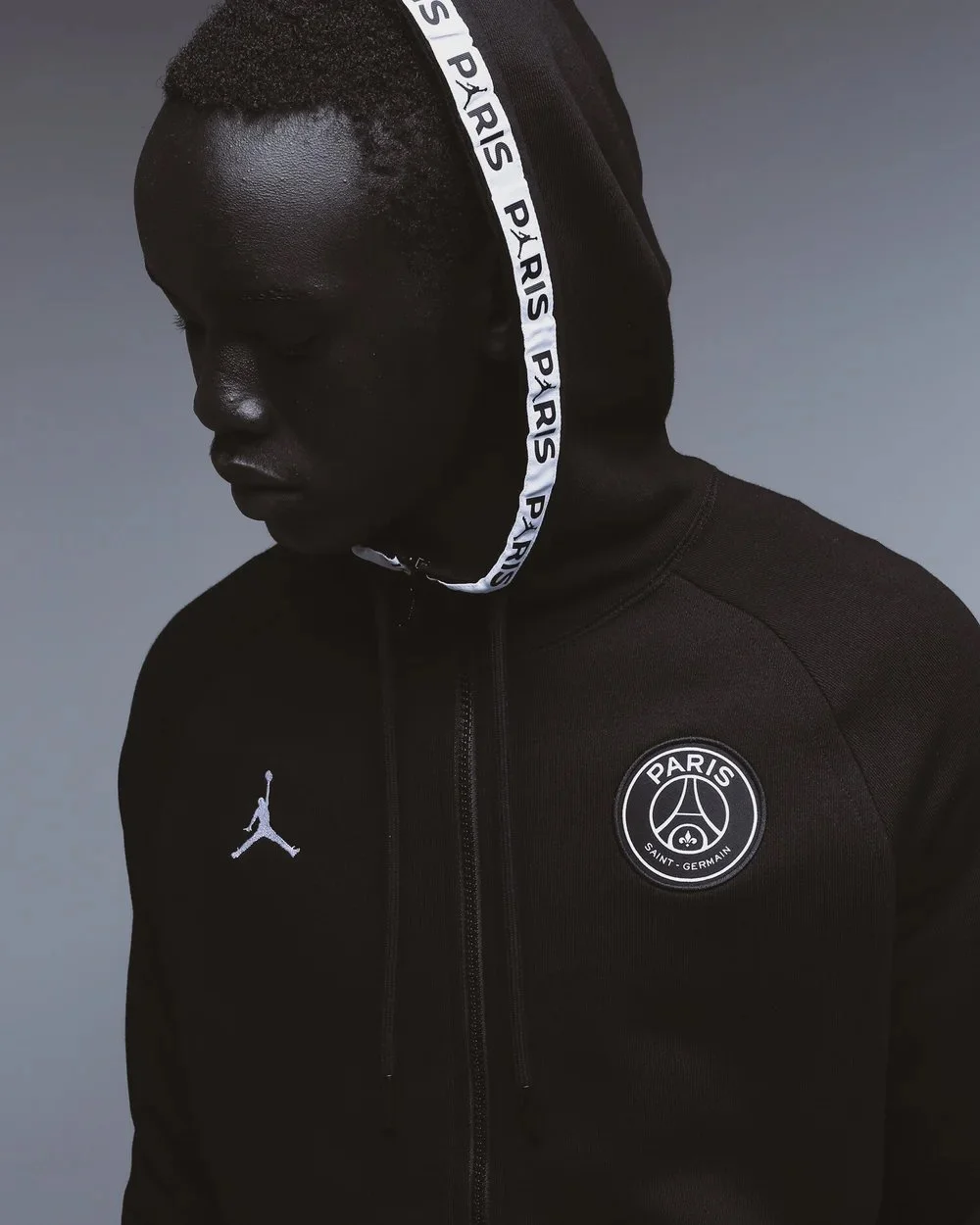 A young man with dark skin wearing a black Paris Saint-Germain hoodie with a white logo on the left chest, and a Jordan brand logo on the right chest. The hoodie has a hood with white drawstrings labeled 'PARIS'. The man is looking downward against a