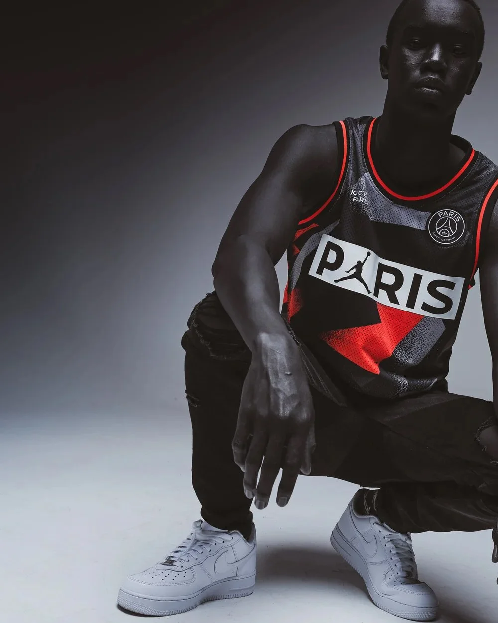 A young man pose in a crouching position wearing a black and red Paris basketball jersey, black pants, and white Nike sneakers. The background is plain and gradient from dark to light.