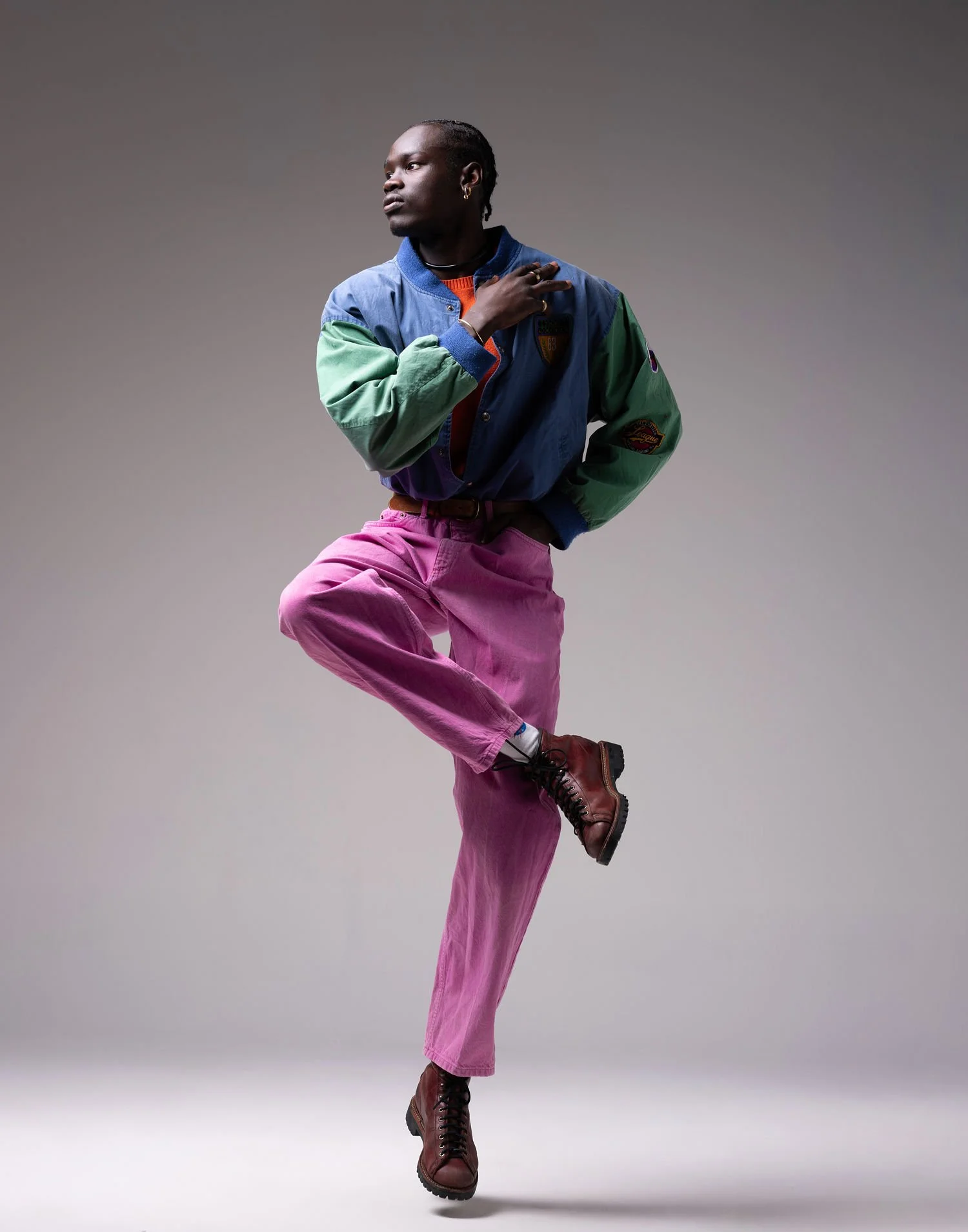 A person with dark skin and braided hair, dressed in colorful clothing, striking a pose against a plain gray background.