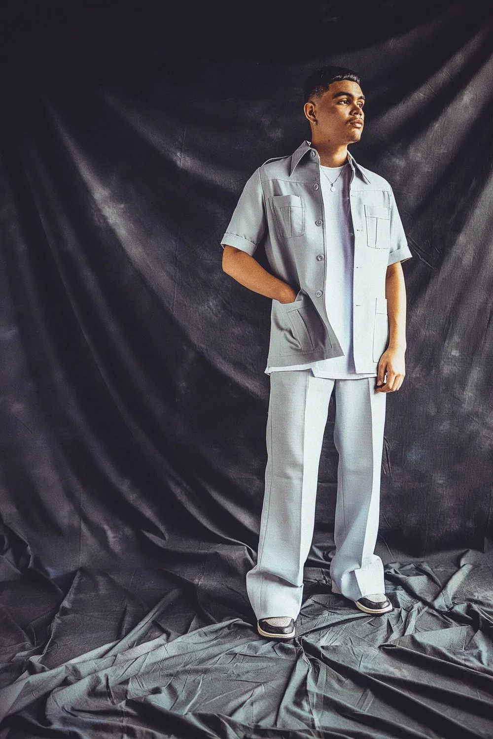A young man standing with hands in pockets, wearing a light grey jacket and matching pants, against a dark textured backdrop.